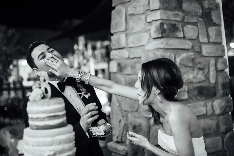 A bride playfully slaps a groom across the face at their wedding reception, with a wedding cake in the foreground.