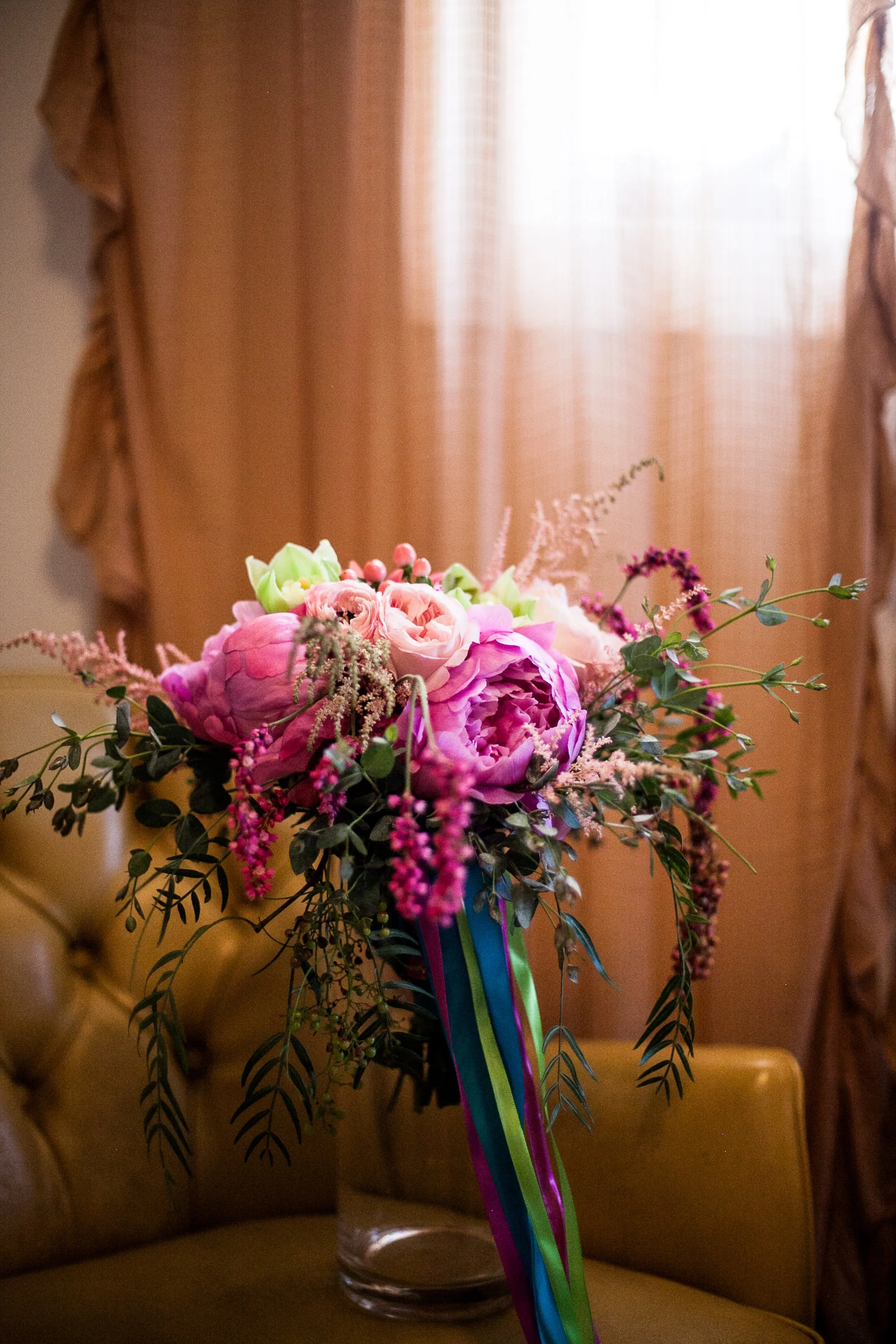 A bouquet of pink and purple flowers with greenery and colorful ribbons in a glass vase on a yellow chair, background with curtains and soft lighting.