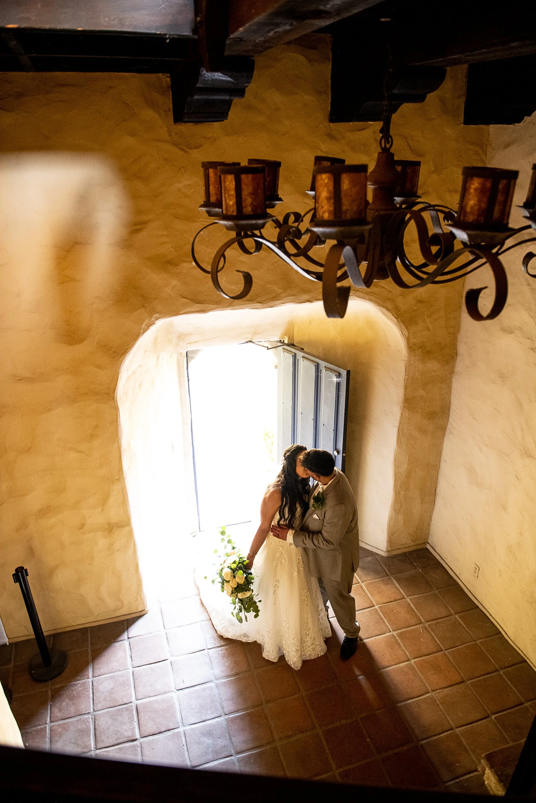 A bride and groom sharing a kiss in front of a window with bright sunlight, inside a rustic room with textured beige walls, tiled floor, and a decorative ceiling lamp overhead.