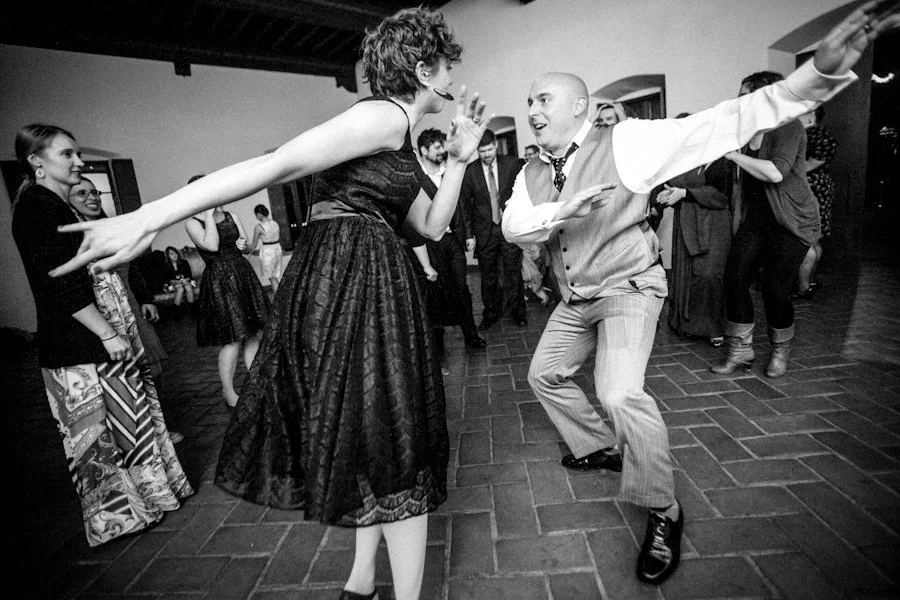 People dancing at a party with a woman in a black dress and a man in a vest and tie energetically dancing.