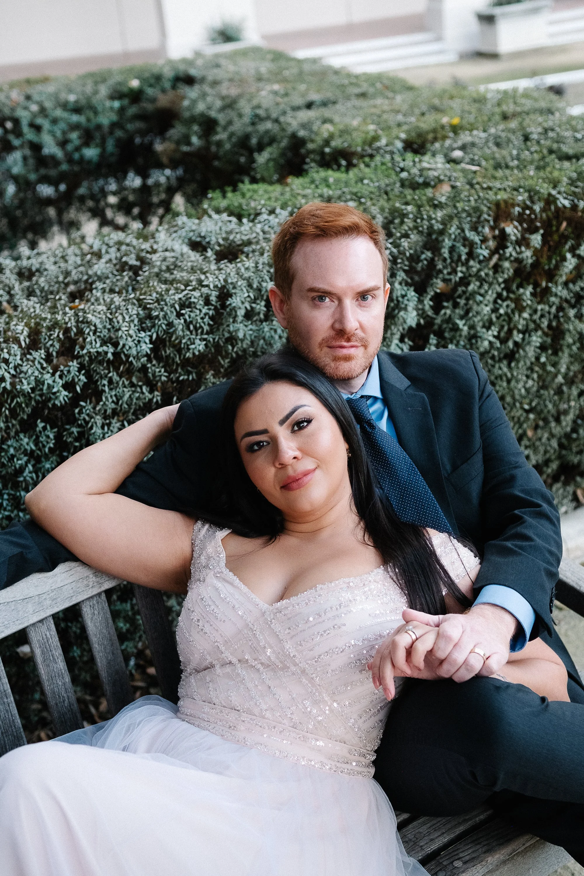 A man and woman sitting closely on a park bench, with the woman leaning against the man, both dressed elegantly, with greenery in the background.