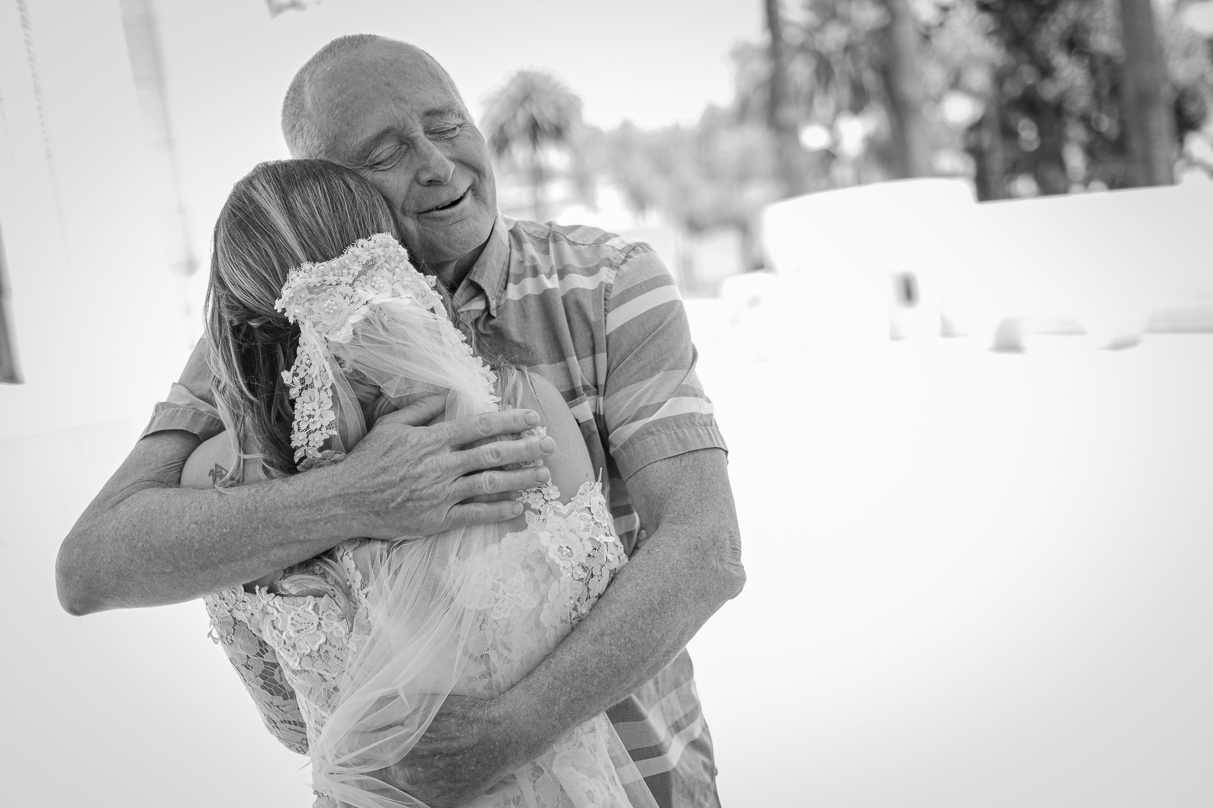 An elderly man hugging a young girl dressed in a wedding veil and dress, sharing a heartfelt moment outdoors.