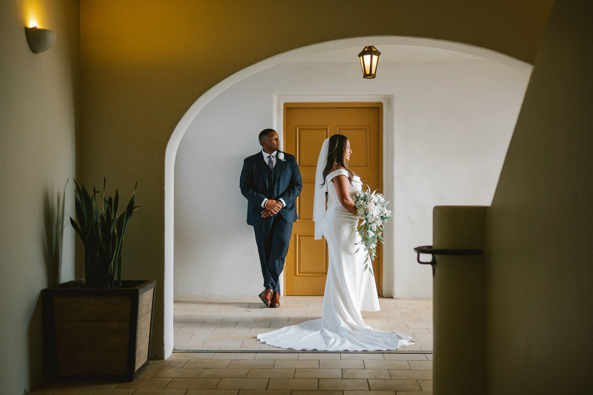 A bride in a white wedding dress holding a bouquet stands in front of a groom in a dark suit, both looking away, in an indoor setting with archways and warm lighting.