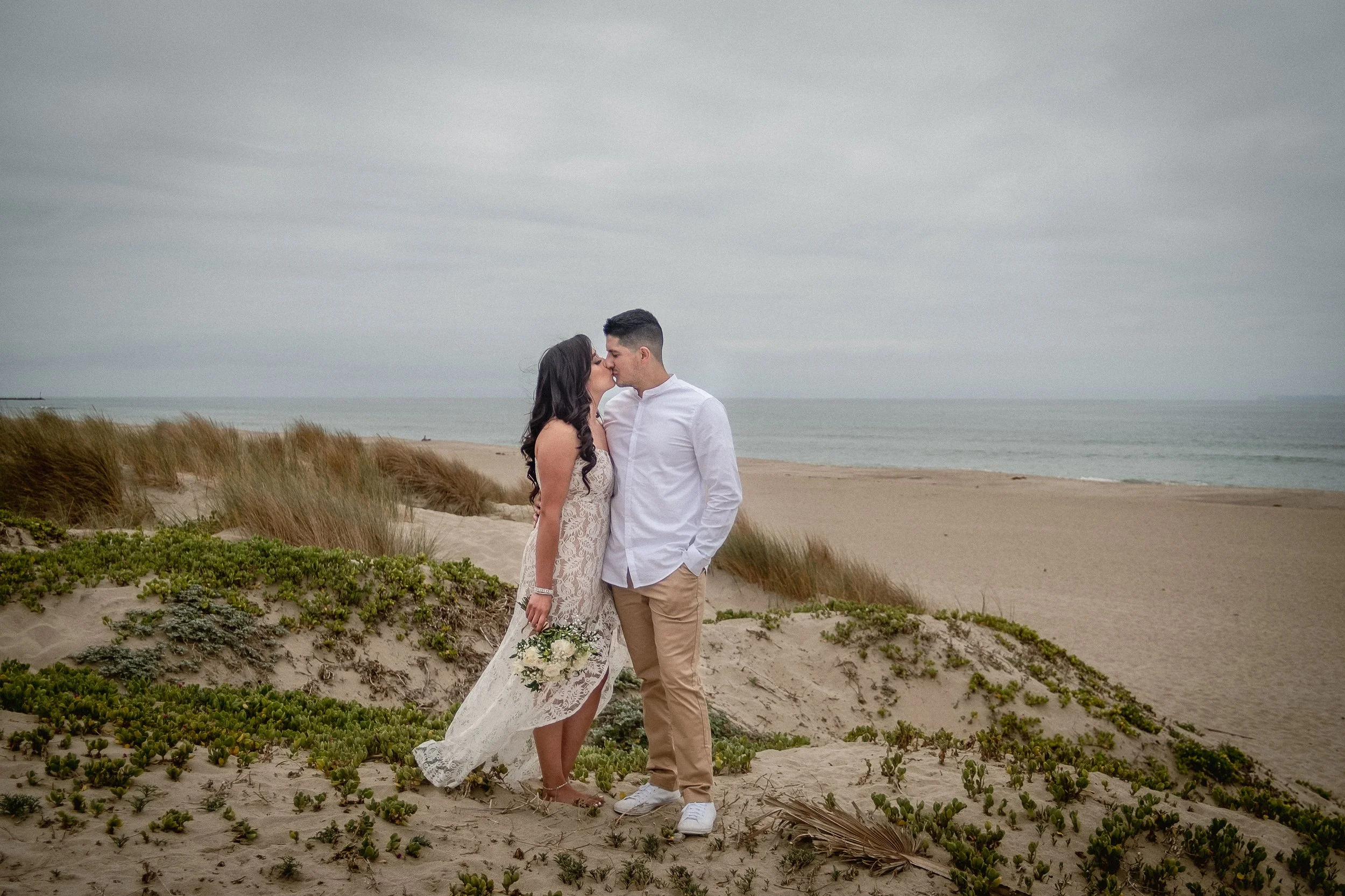 A bride and groom sharing a kiss on a sandy beach with grassy dunes and the ocean in the background.