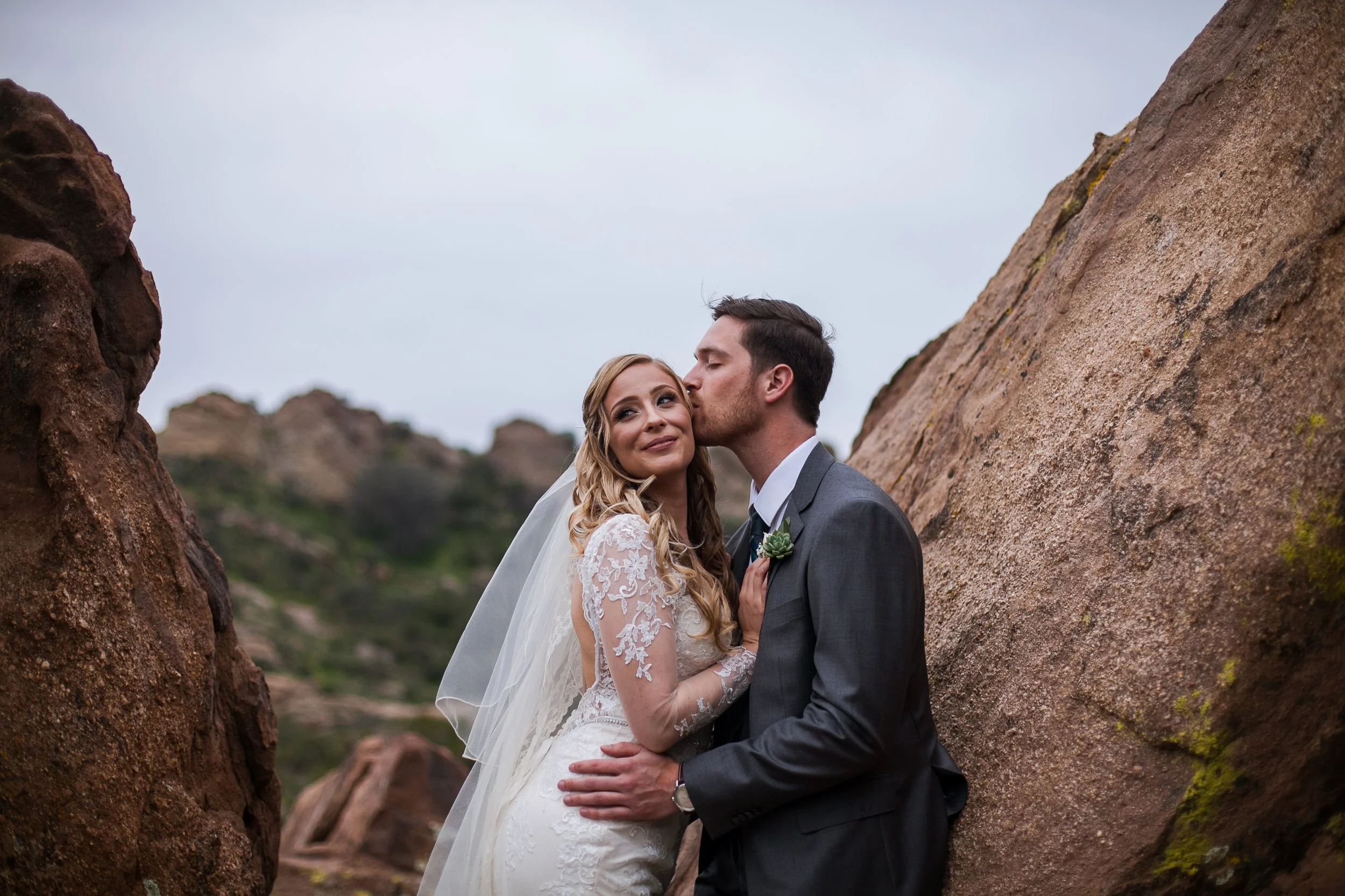 Bride and groom standing between rocks during wedding photoshoot, groom kissing bride's temple, outdoor natural landscape in background.