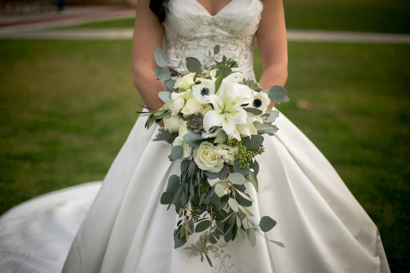 Bride in white wedding gown holding a cascading bouquet of white roses, lilies, anemones, and greenery outdoors.