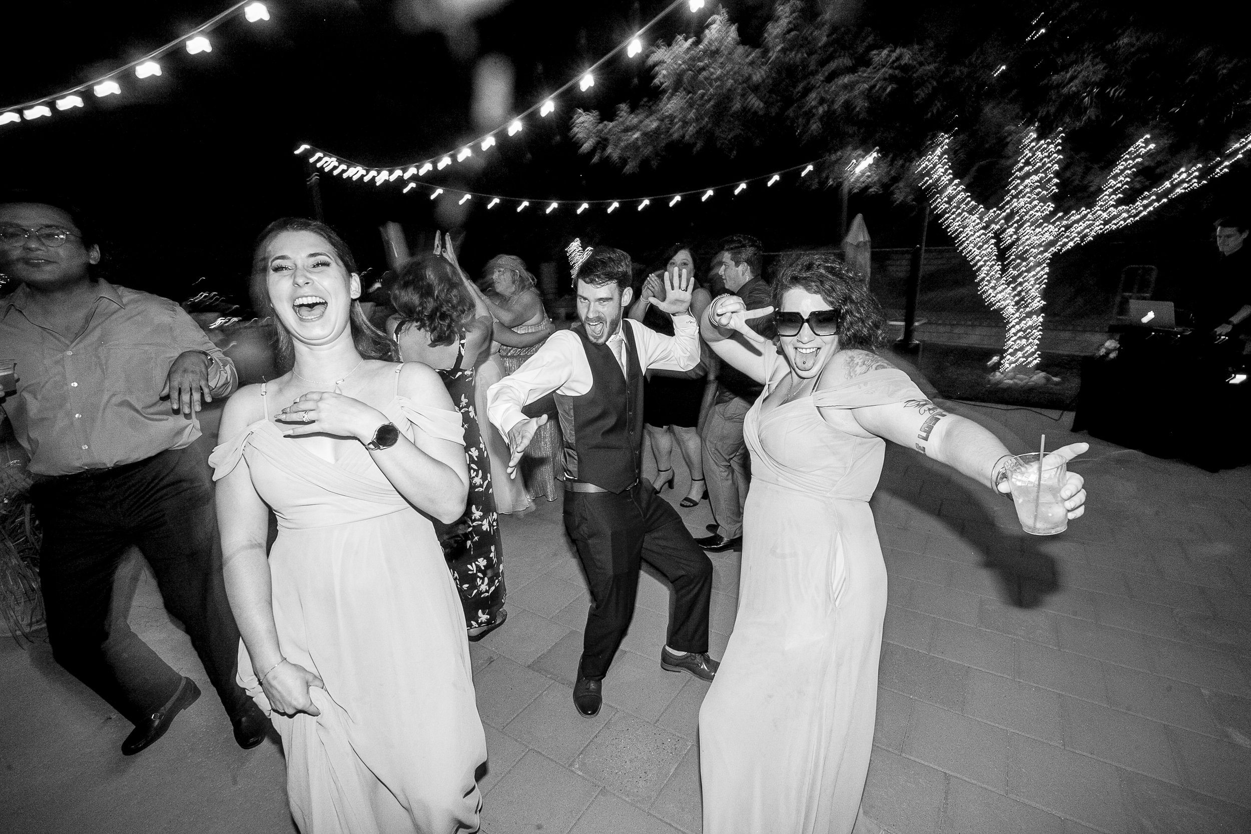 People dancing and celebrating at an outdoor nighttime party with string lights and decorated trees.