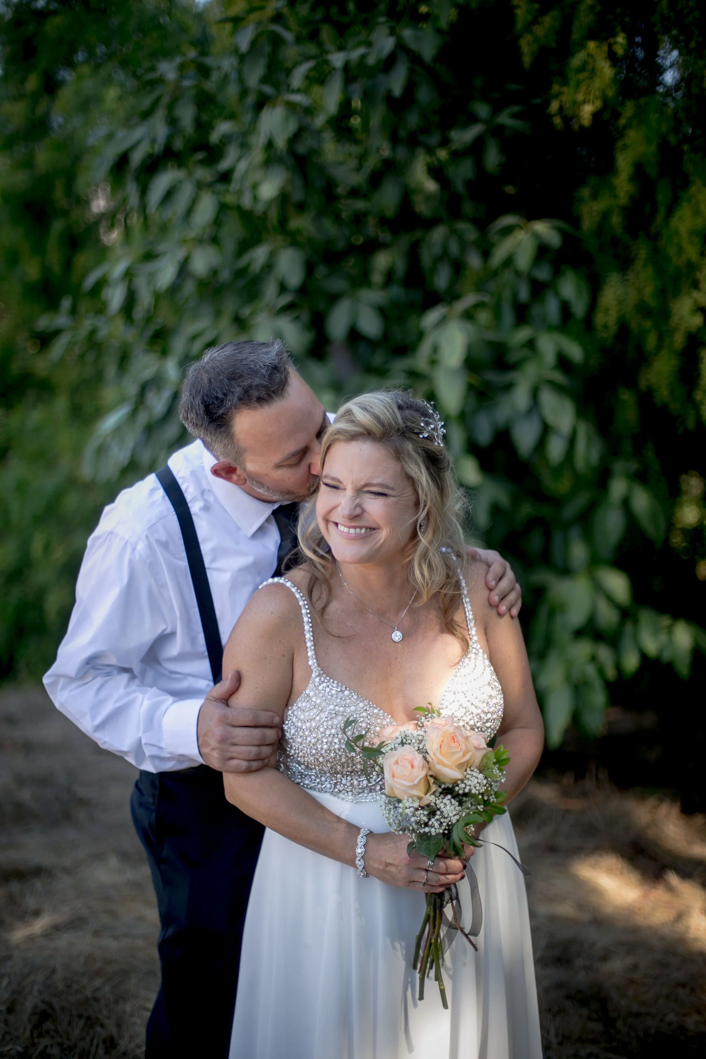 A bride holding a bouquet of pink roses and white baby's breath, smiling with eyes closed, while a groom kisses her on the head outdoors amidst greenery.