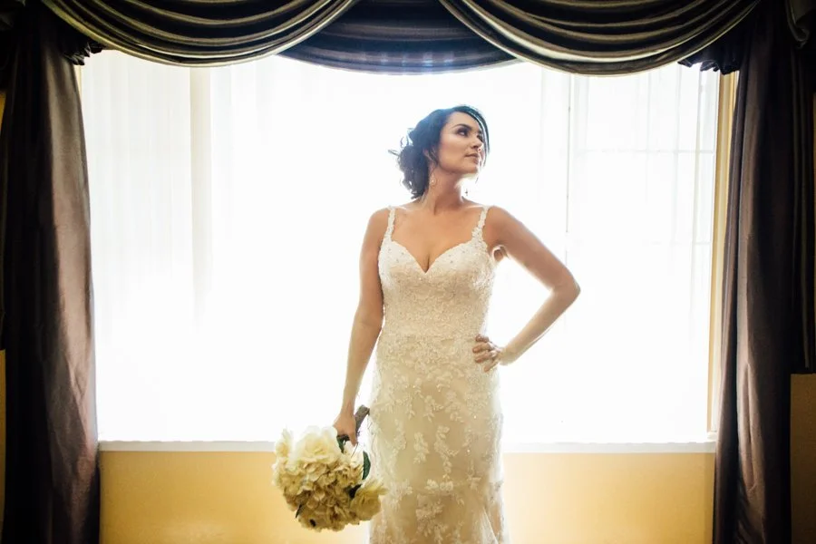 Bride in a lace wedding dress holding a bouquet, standing in front of a window with white curtains and dark drapes.