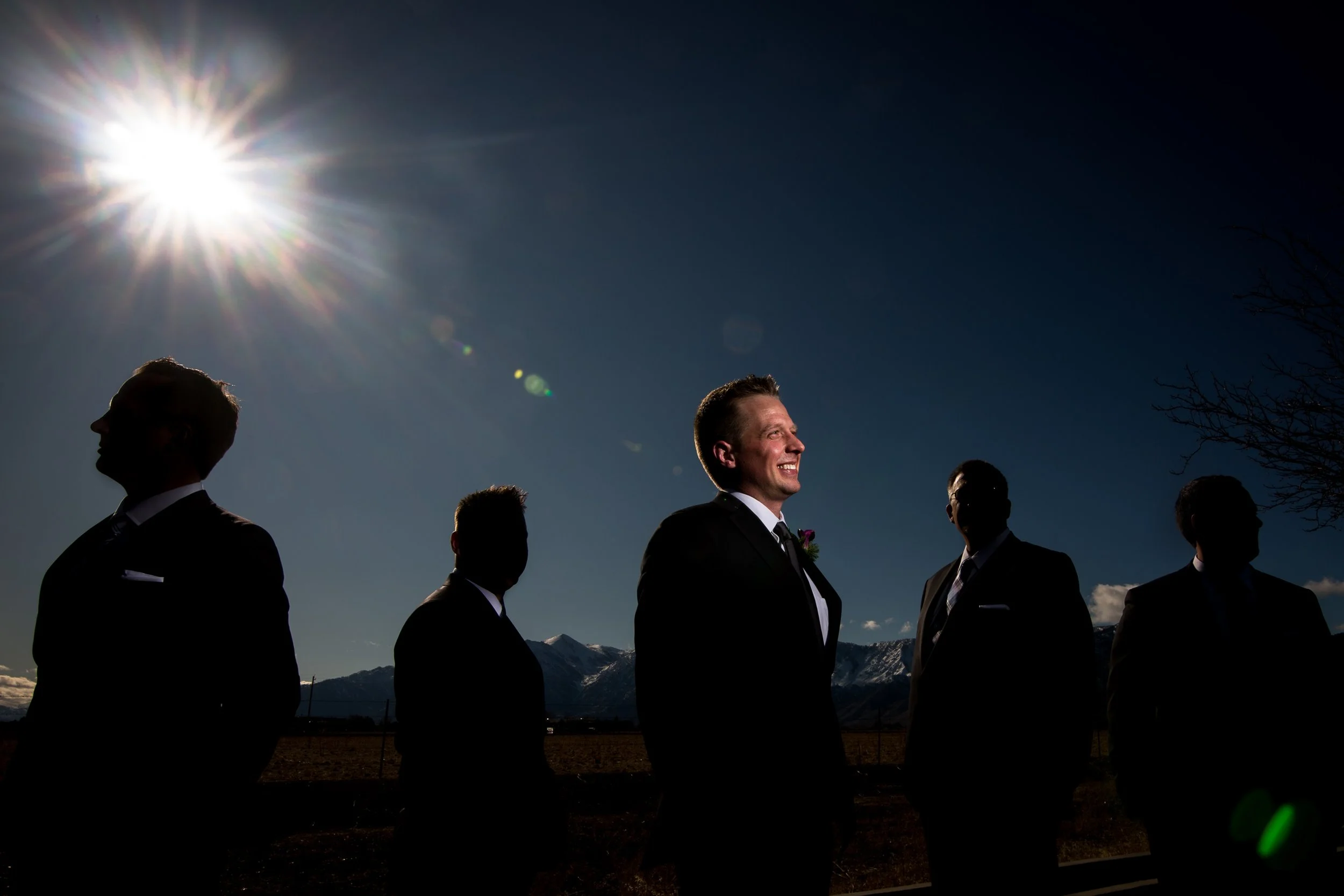 A group of five men dressed in suits standing outdoors with mountains in the background, sunlight shining overhead, and one man smiling in the center.