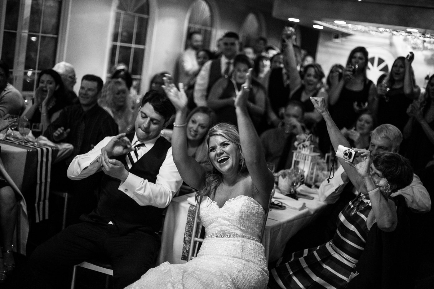 Black and white photo of a wedding reception with guests, including a woman in a wedding dress smiling and raising her arms, and a man in a suit sitting beside her. Other guests are clapping and taking photos, watching the celebration.