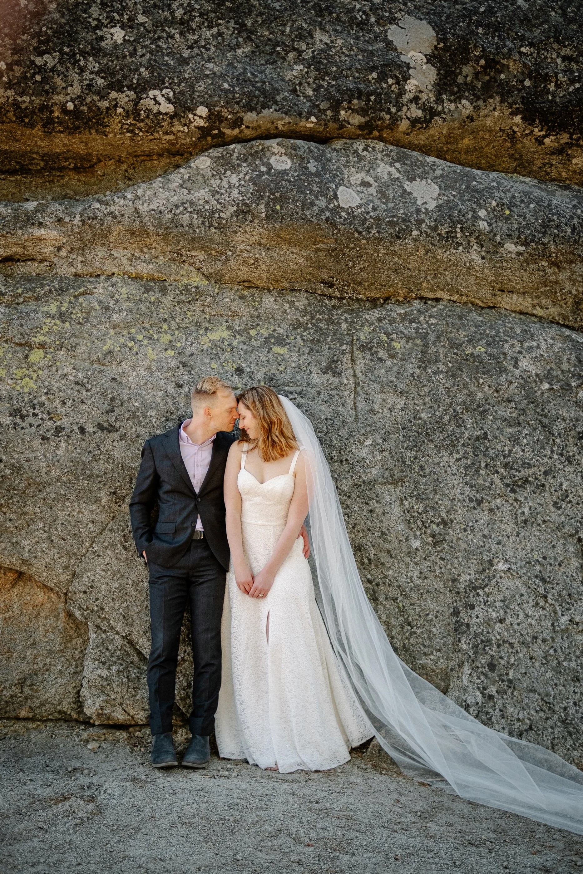 A bride and groom standing close together with foreheads touching against a large rock wall, outdoors during their wedding.