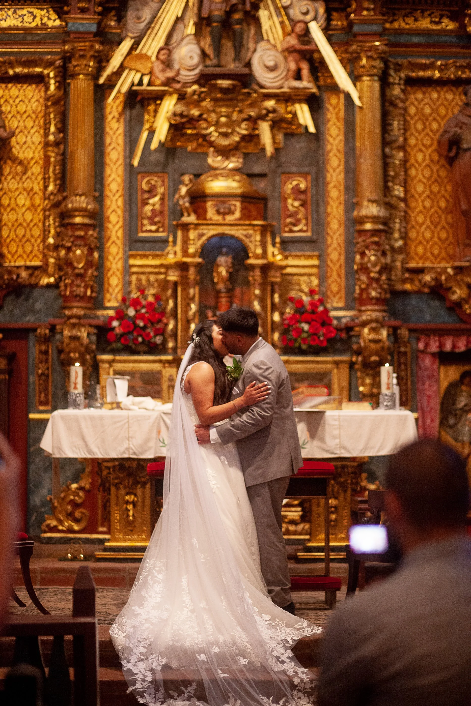 A newlywed couple sharing a kiss during their wedding ceremony inside a church with ornate gold and wood decorations and red floral arrangements in the background.