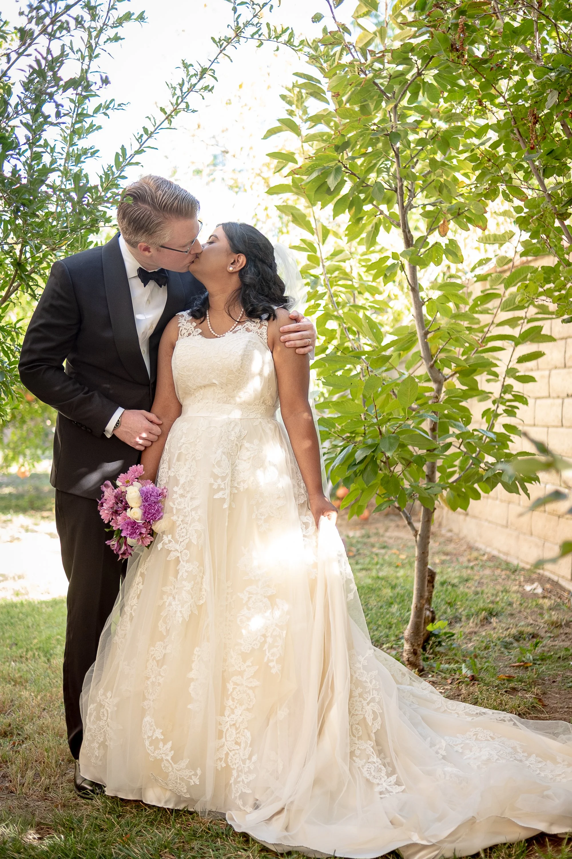 A bride and groom kissing outdoors, the bride holding a bouquet of purple and white flowers, surrounded by green trees.