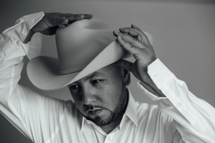A man wearing a white dress shirt puts on a light-colored cowboy hat, adjusting it with both hands, against a plain background.