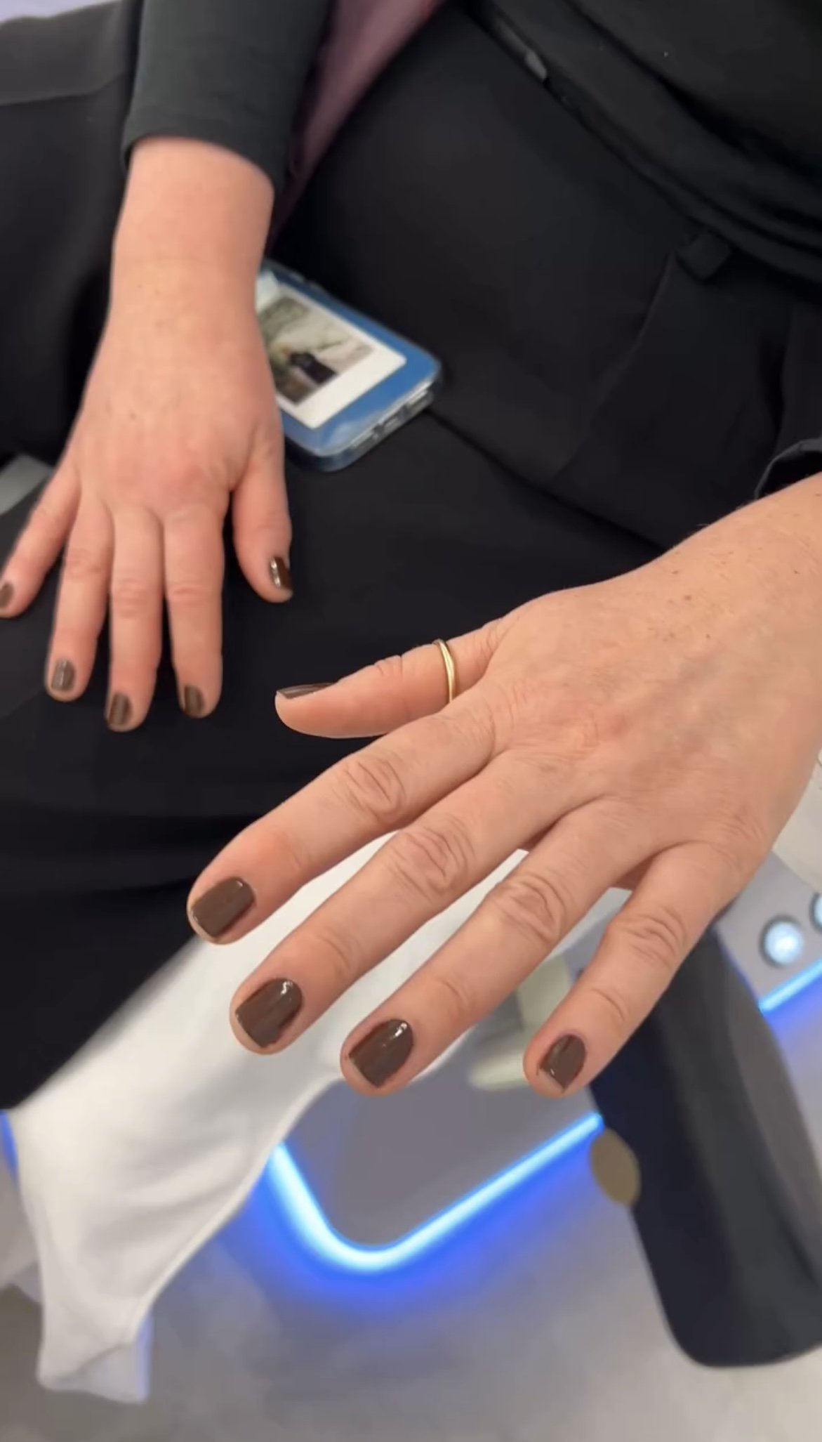 Close-up of a person's hands with brown nail polish and a gold ring, resting on a black fabric with a cellphone in the background.