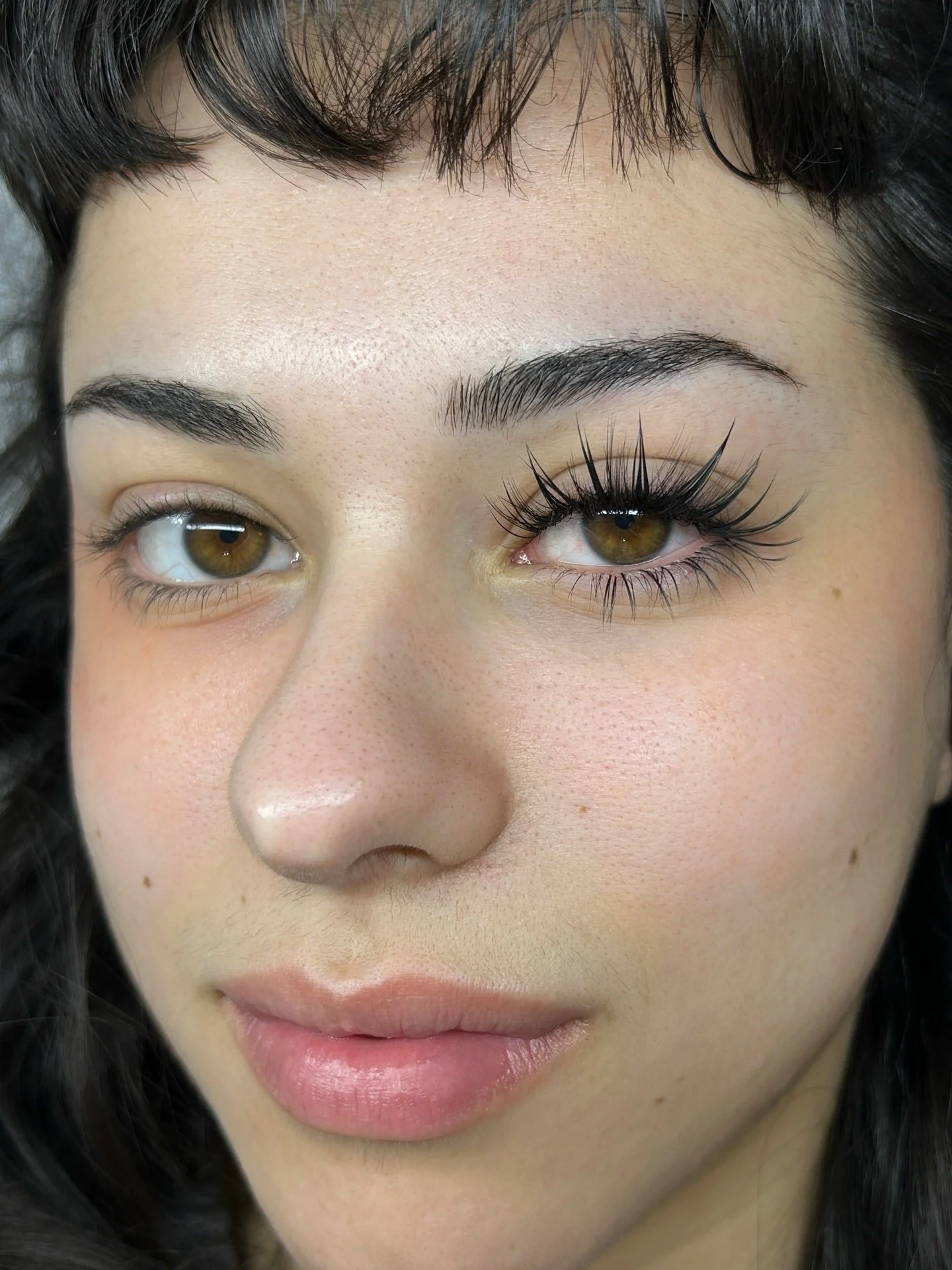 Close-up of a woman’s face showing brown eyes, black hair, and soft makeup, featuring Medusa-style eyelash extensions with bold wispy spikes and defined lash texture. Focus on dramatic spiky lashes around the eyes.