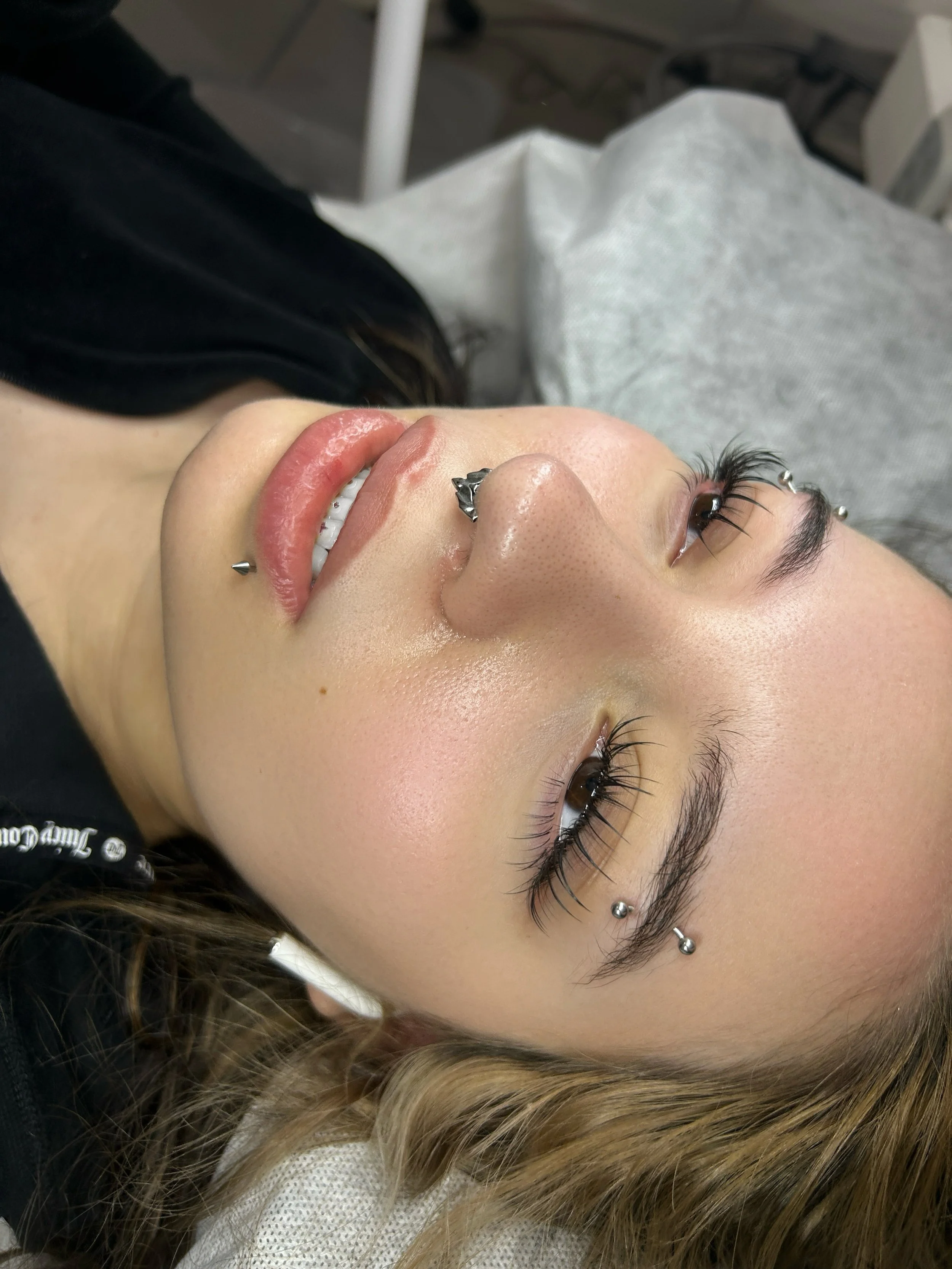 A woman with multiple facial piercings, including on her nose and ears, lying down with her eyes open, smiling slightly, with long dark hair and bright lighting.