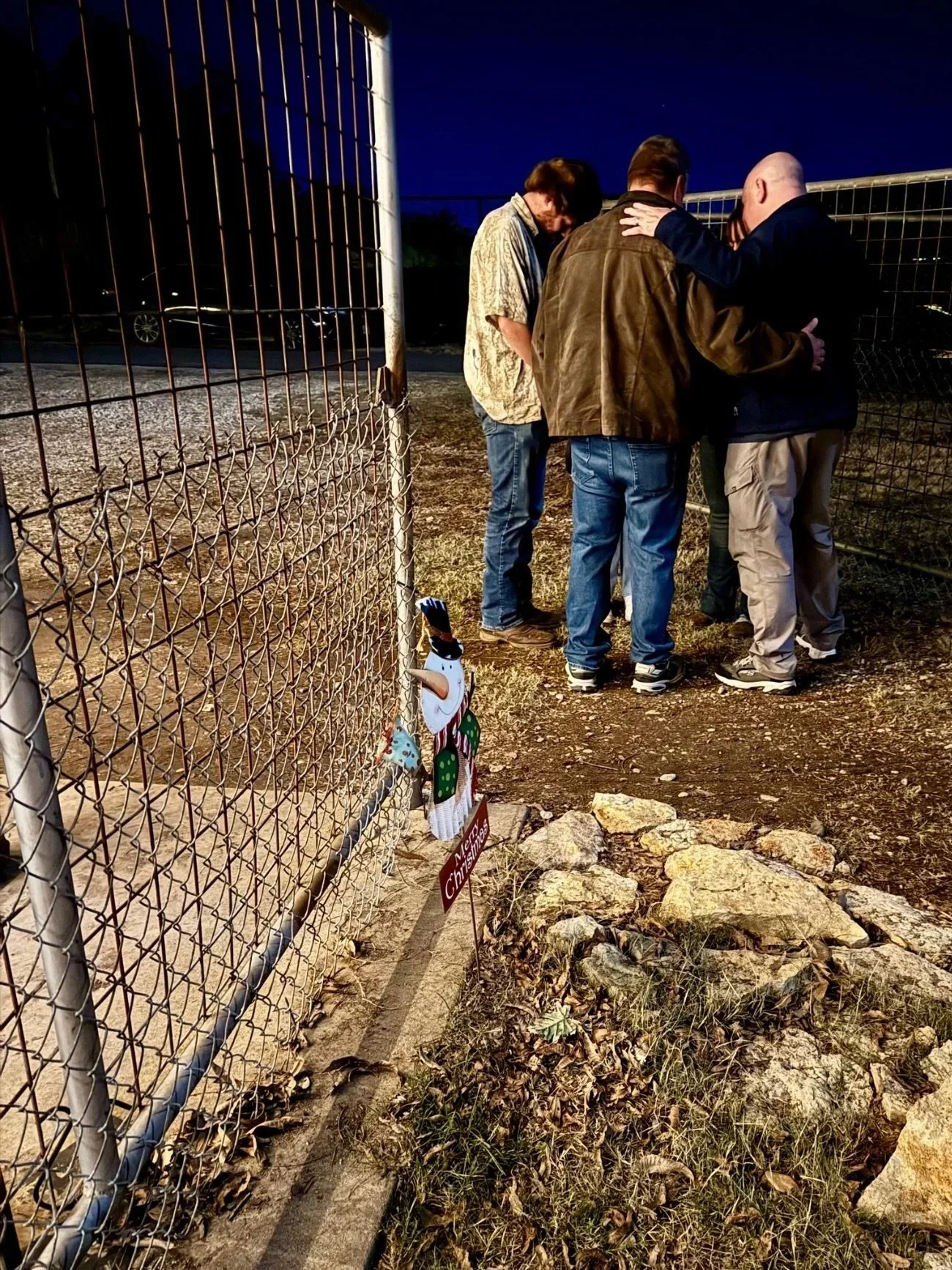 Four men standing close together, engaged in prayer or a moment of silence, behind a chain-link fence with a Christmas decoration of a snowman attached to it, on a dirt ground with rocks, during evening or night.