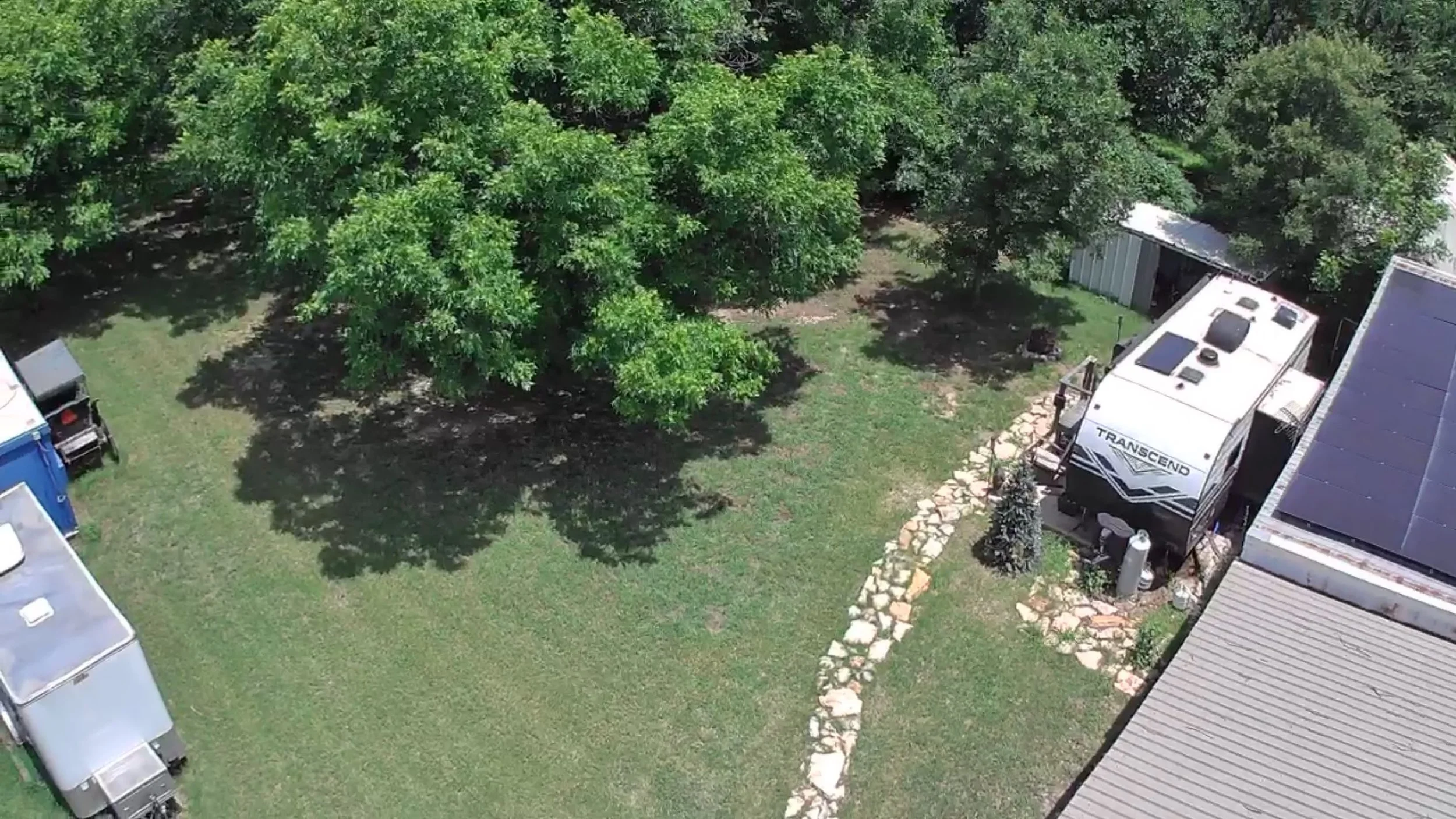 An aerial view of a backyard with lush green trees, a stone pathway, and a white RV parked next to a house with a solar panel roof.