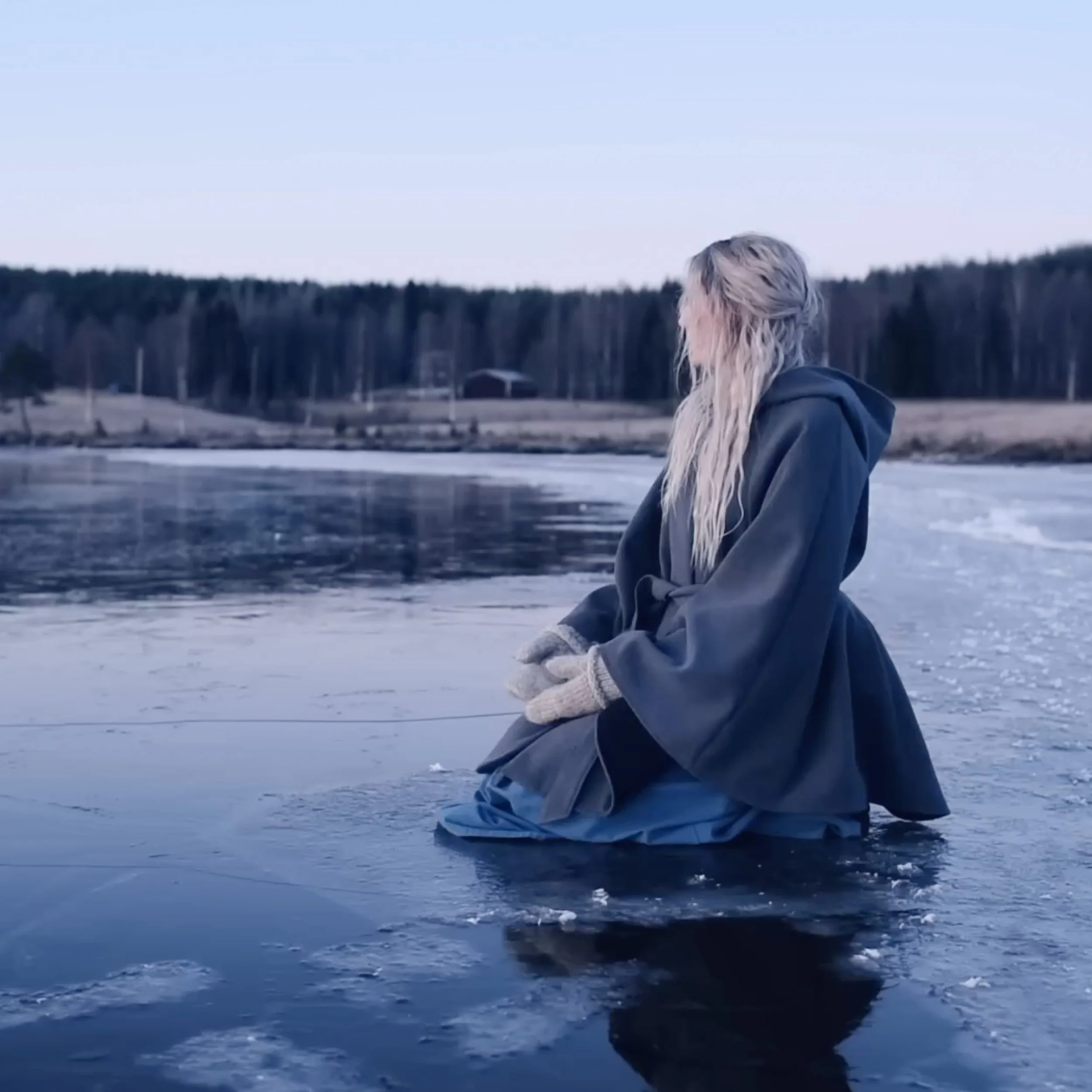 Frau in dunkler Jacke und gestrickten Handschuhen kniet auf gefrorenem Wasser in einer Winterlandschaft, mit Bäumen im Hintergrund.