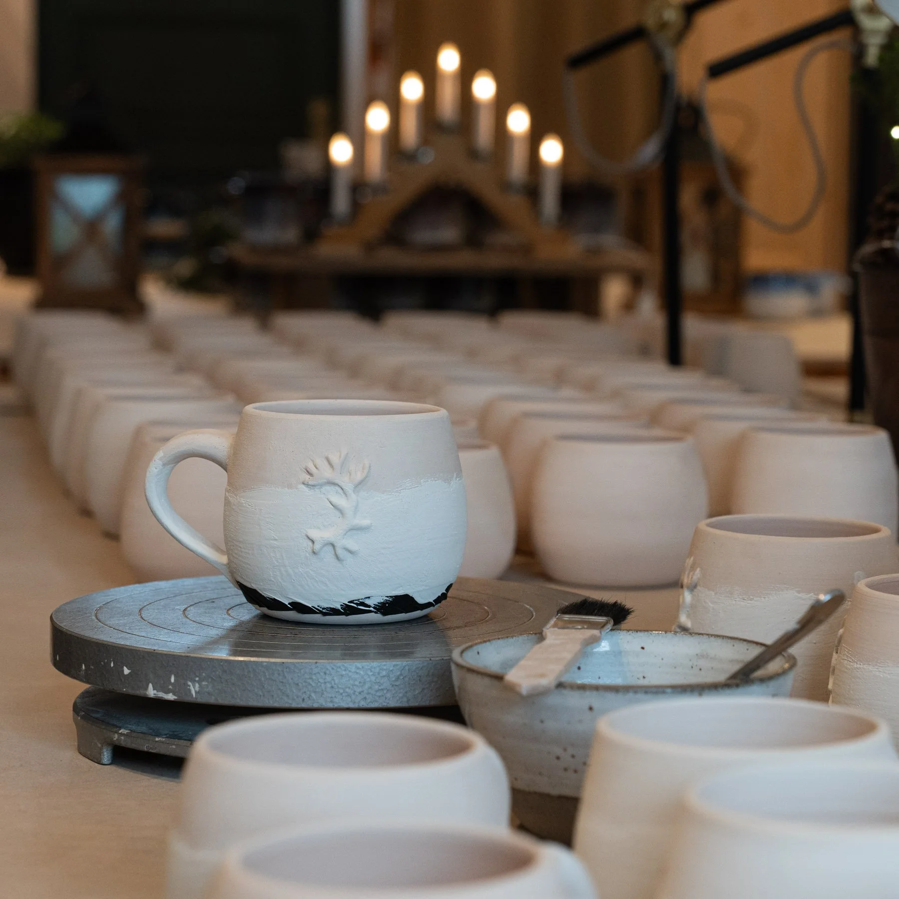 Ungeschliffene Keramikbecher und -schalen in einer Werkstatt, einige sind auf einer Töpferscheibe, im Hintergrund eine Kerzenleiste.