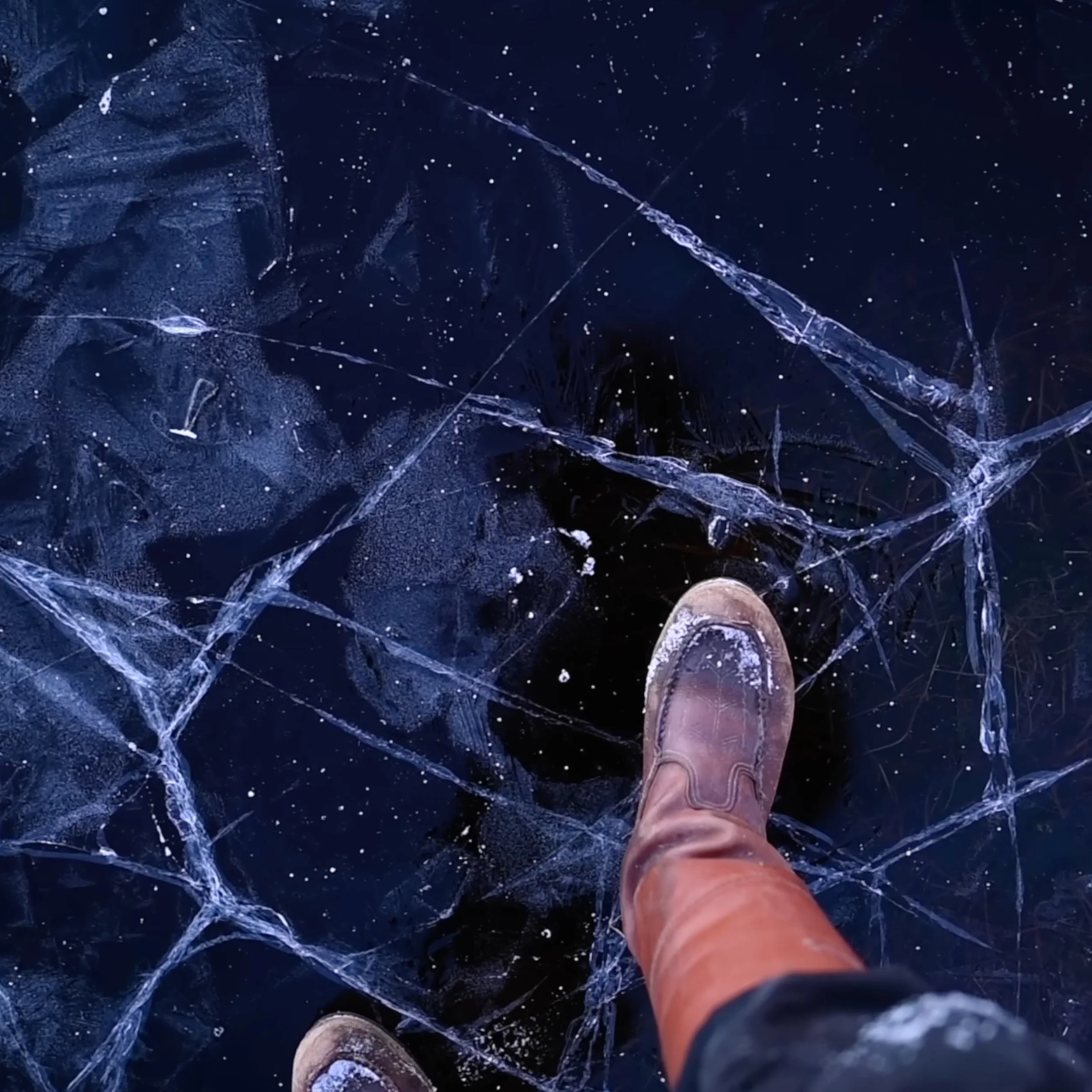 Eine Person steht auf gefrorenem Wasser mit durchsichtigen Eisschichten und Rissen, trägt braune Stiefel und schwarze Hosen.