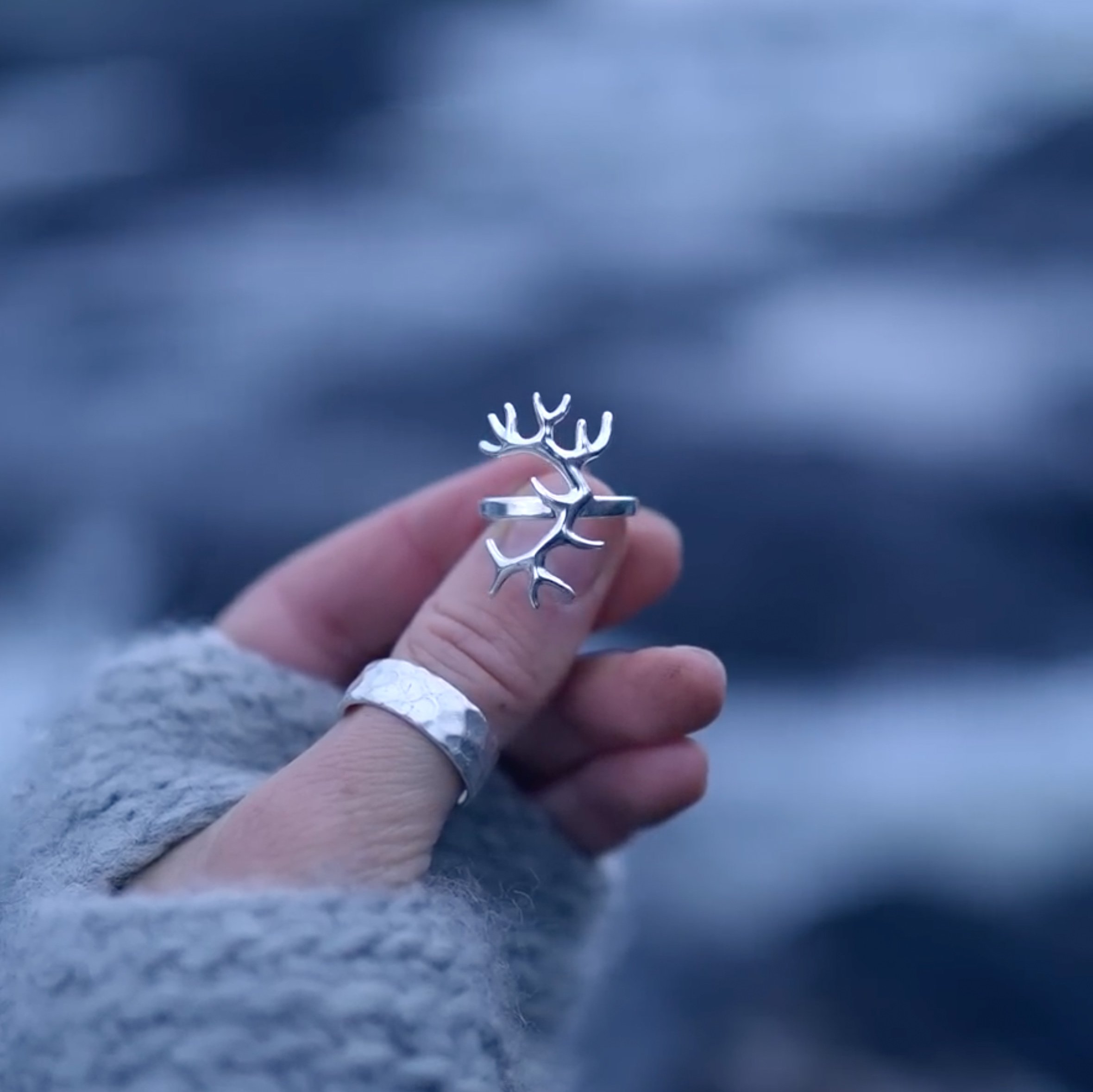 Eine Hand trägt einen Silberring in Form von Hirschgeweih, gehalten vor einem verschwommenen, dunklen Hintergrund.