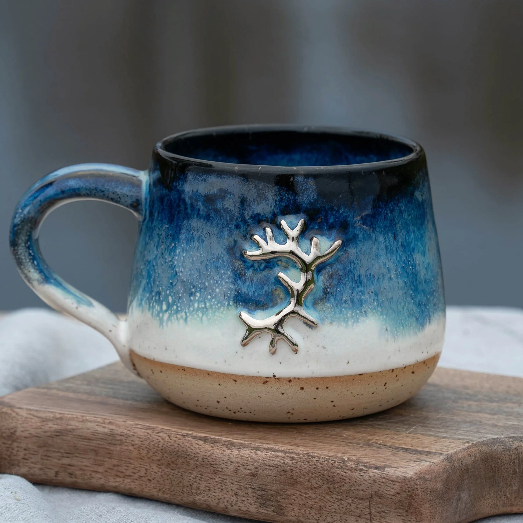 Eine handgefertigte Keramiktasse mit blauer, weißer und cremefarbener Glasur, verziert mit einem silbernen Ast-Design, steht auf einem Holzbrett.