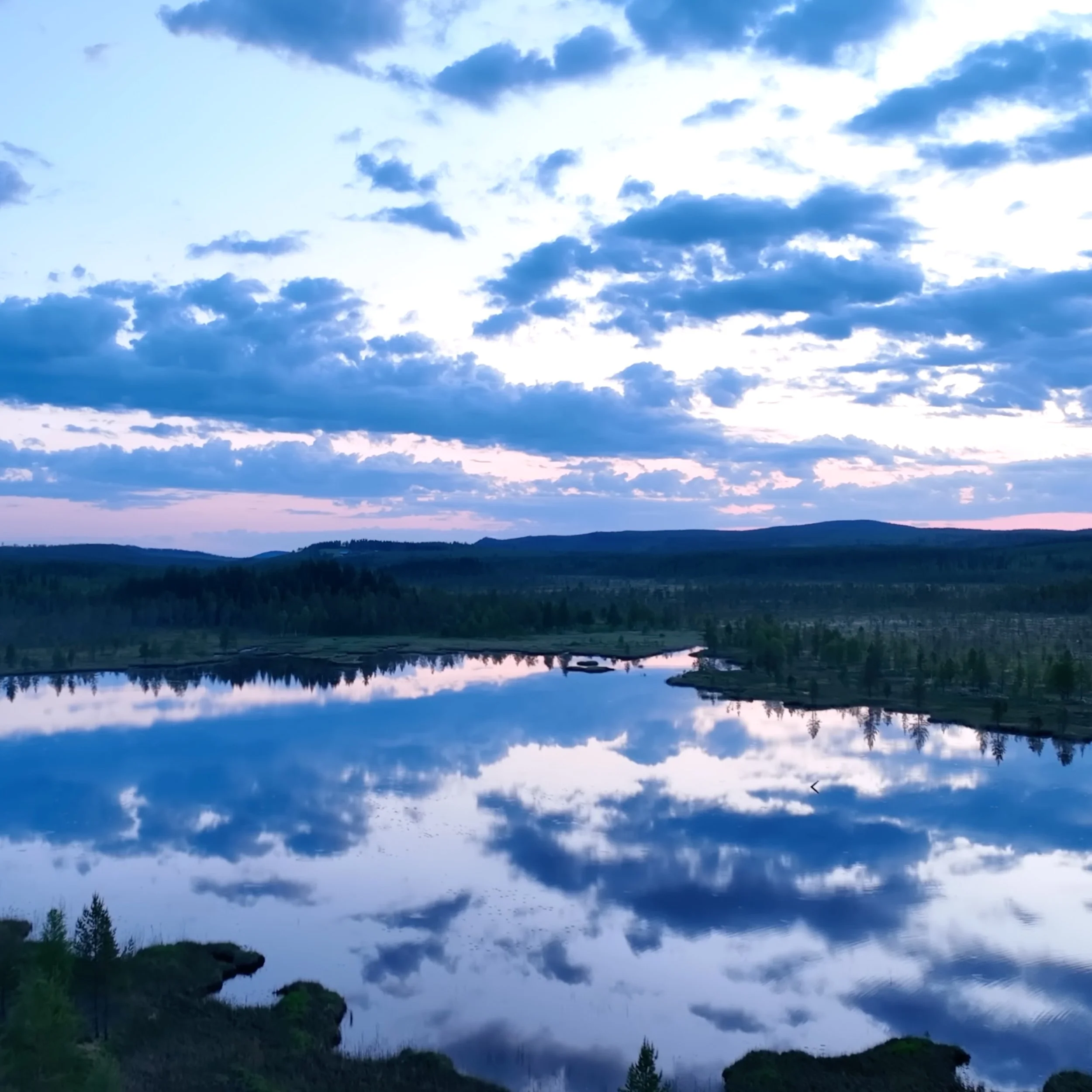 Ein ruhiger See spiegelt den Himmel mit Wolken während des Sonnenuntergangs oder Sonnenaufgangs wider, umgeben von Wäldern und sanften Hügeln.