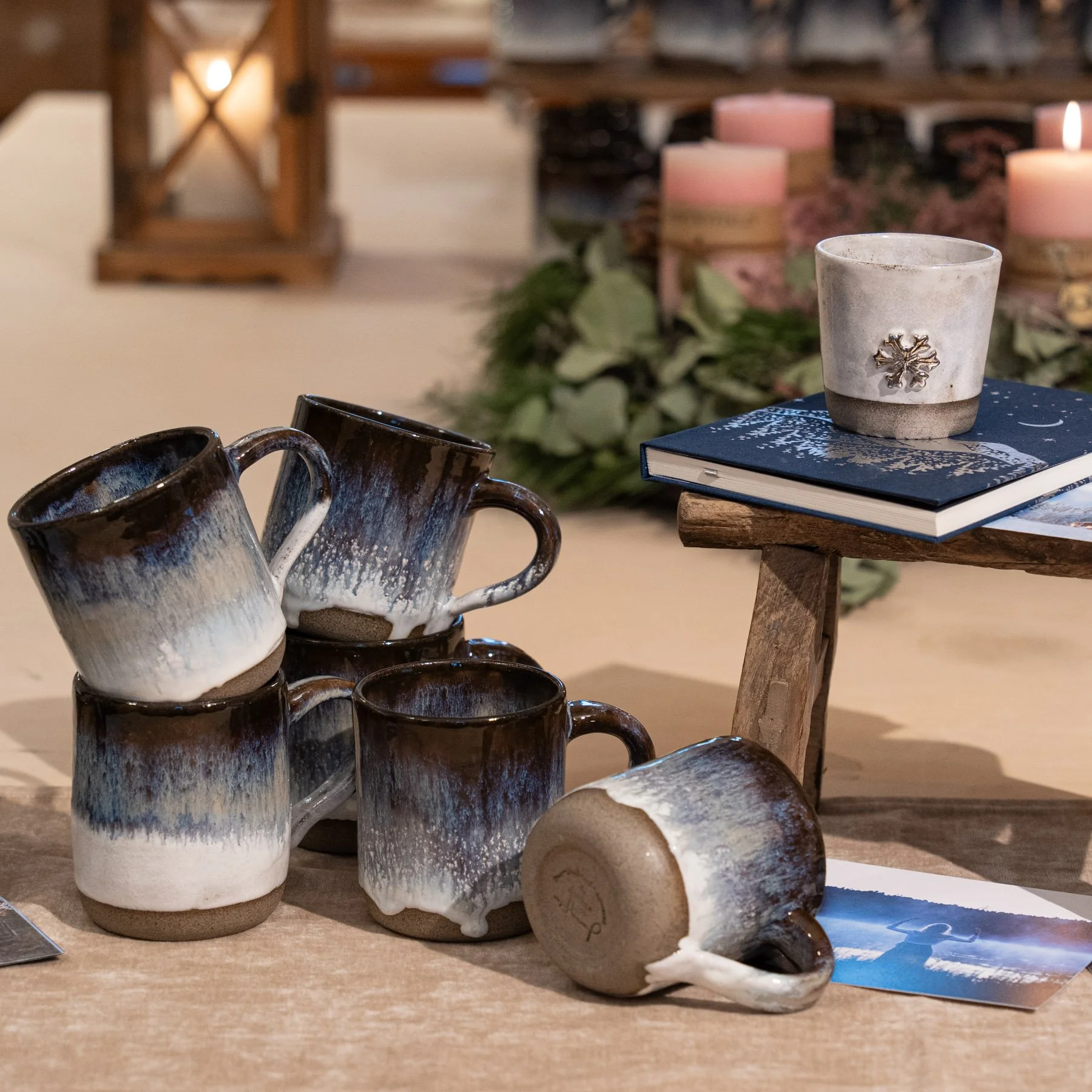 Mehrere handgefertigte Keramiktassen in einem braunen Farbton mit weißer Überläufe, einige stehen auf einem Holztisch, eine liegt umgedreht. Im Hintergrund sind Kerzen, grüne Zweige und ein Buch sichtbar.