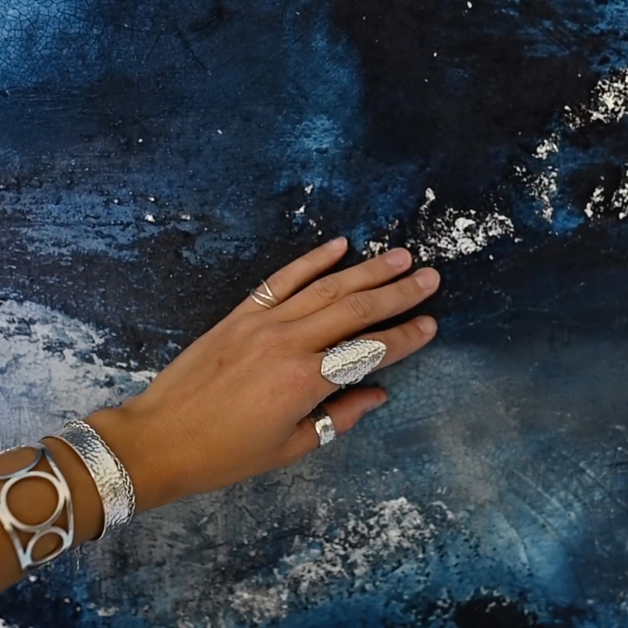 Eine Hand mit mehreren silbernen Ringen und einem großen silbernen Ring, die auf eine dunkelblaue, strukturierte Oberfläche gelegt ist.
