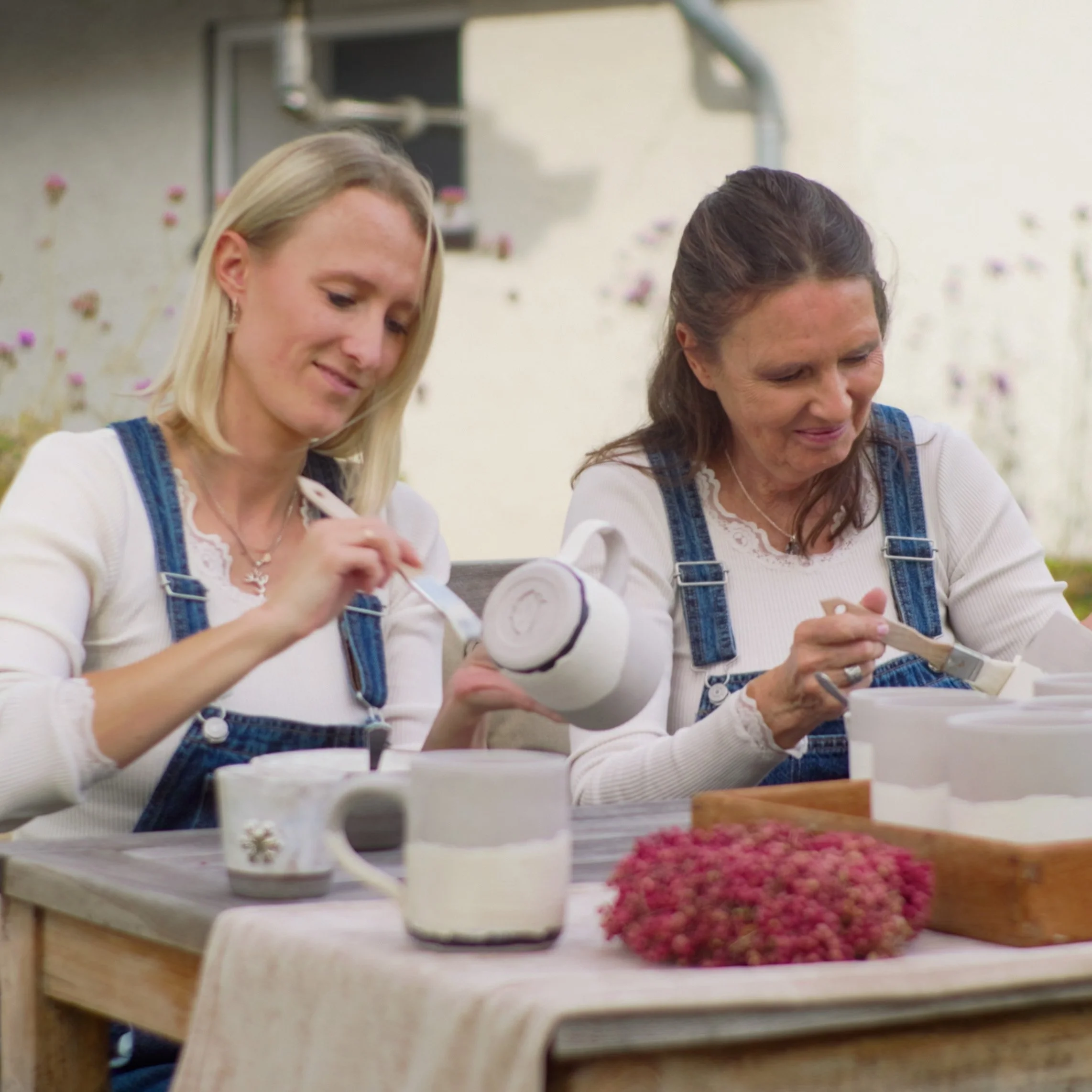 Zwei Frauen beim Malen und Basteln im Garten, sitzend an einem Holztisch mit Tassen und Bastelmaterialien, im Hintergrund Blumen und eine weiße Hauswand.