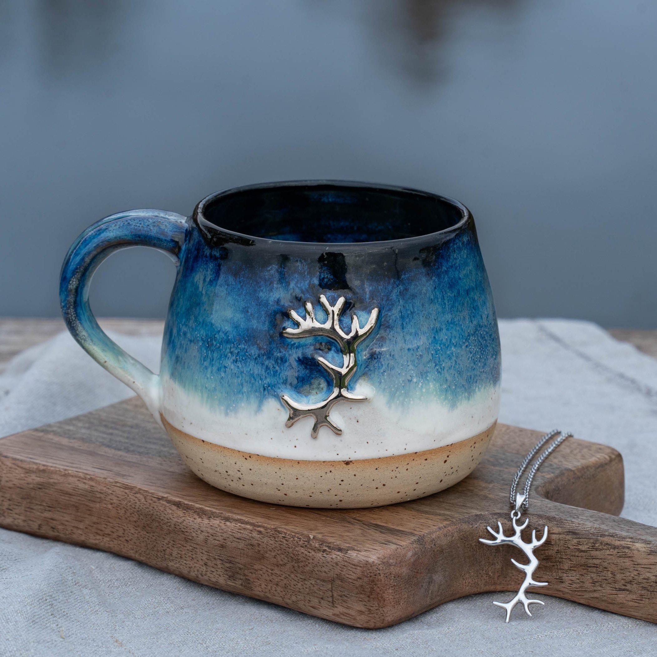 Eine bunte Tasse mit blauer und weißer Glasur und einem silbernen Baum-Dekor, auf einem Holzbrett mit einer passenden Kette antiker Baum-Anhänger, vor einem grauen Hintergrund.