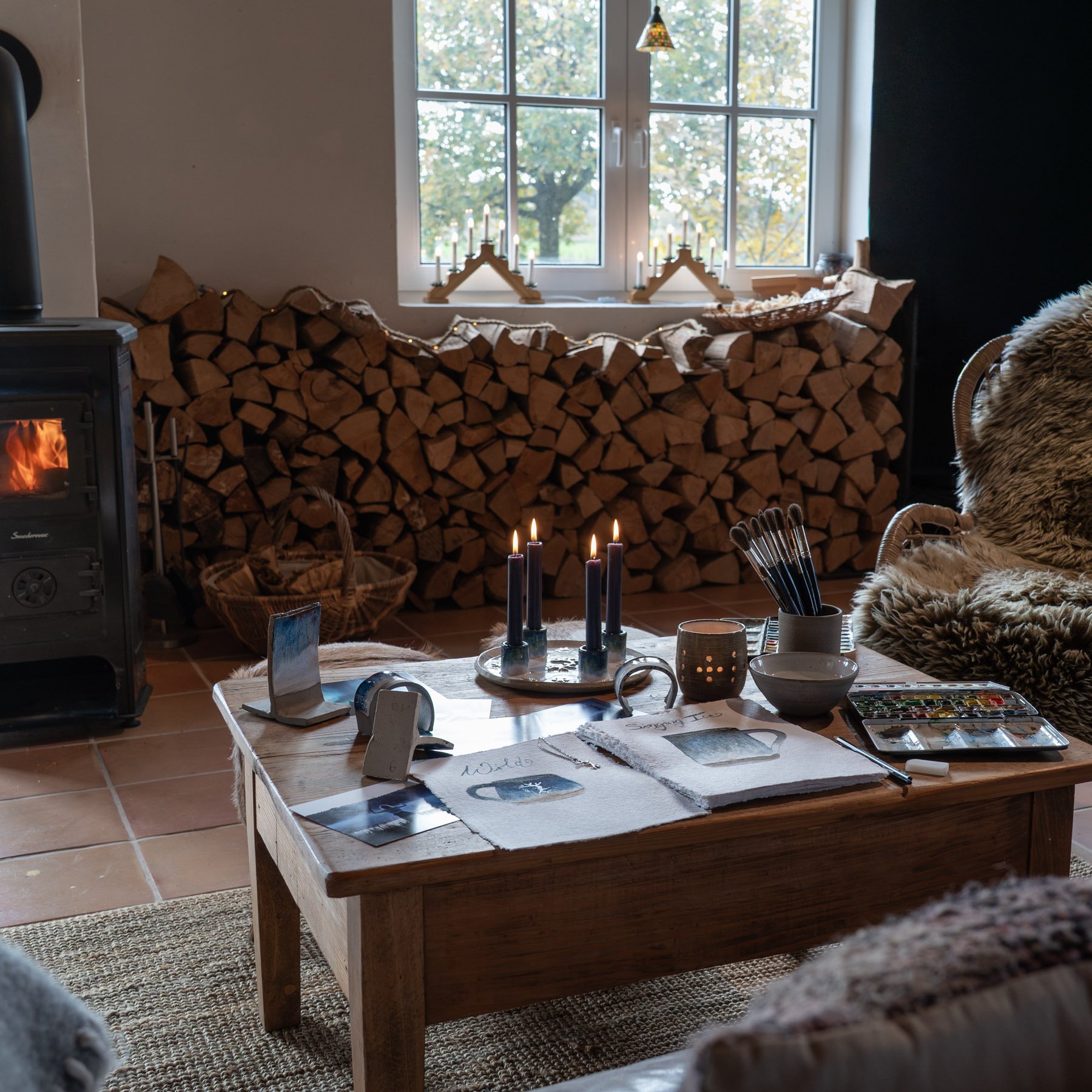 Ein gemütliches Wohnzimmer mit Kamin, Holzstapel im Hintergrund, einer Kaffeetafel mit Kerzen, Malutensilien, Fotokarten und einem Sofa mit Fell.