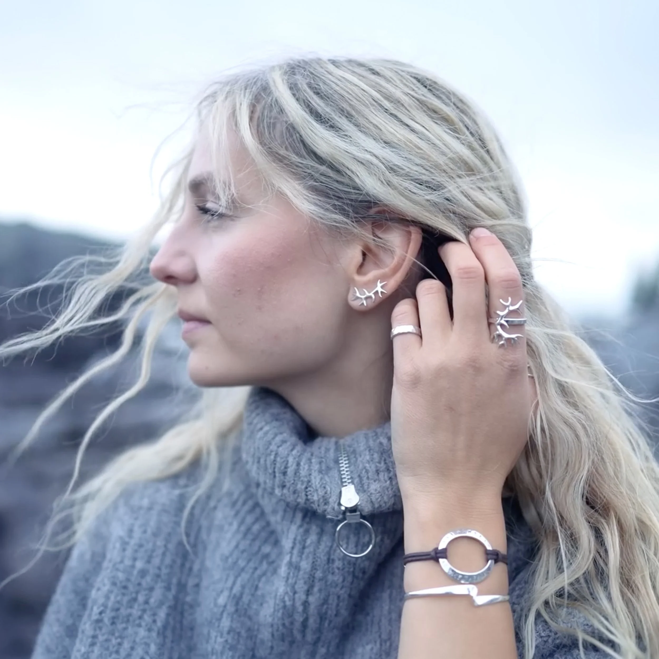 Nahaufnahme einer Frau mit blondem, gewelltem Haar, die silberne Ohrringe, Ringe und Armbänder trägt, während sie an einer Seite ihres Kopfes berührt.