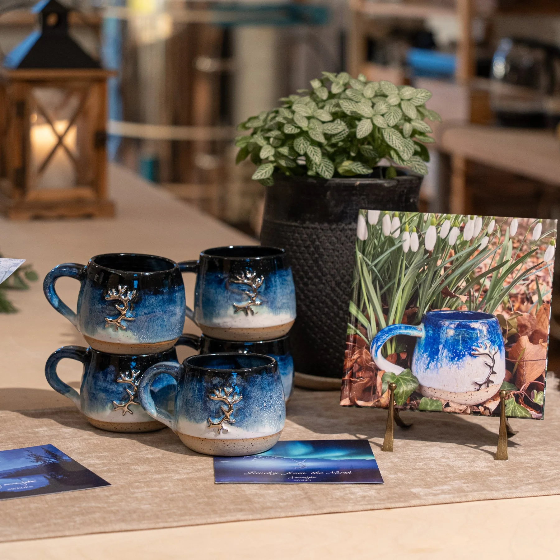 Vier handgefertigte Keramik-Tassen mit einem Baum-Design in Blau und Weiß, eine Pflanze in einer schwarzen Vase, eine kleine Leinwand mit einem ähnlichen Tassen-Motiv und some Informationen auf Karten auf einem Holz- und Stofftisch.