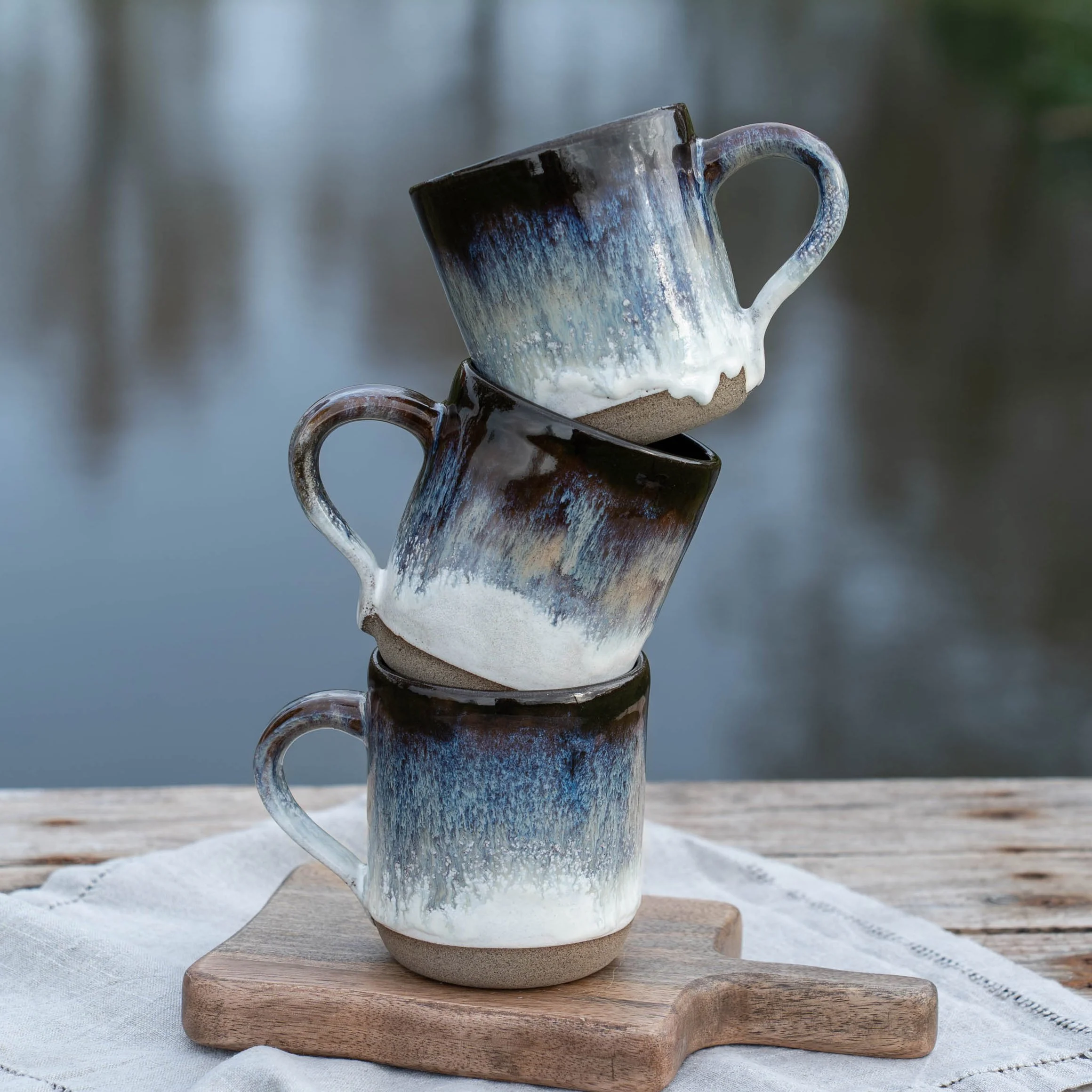 Drei keramikbecher mit künstlerischem Farbdesign, gestapelt auf einem Holzbrett vor einer Wasserlandschaft.