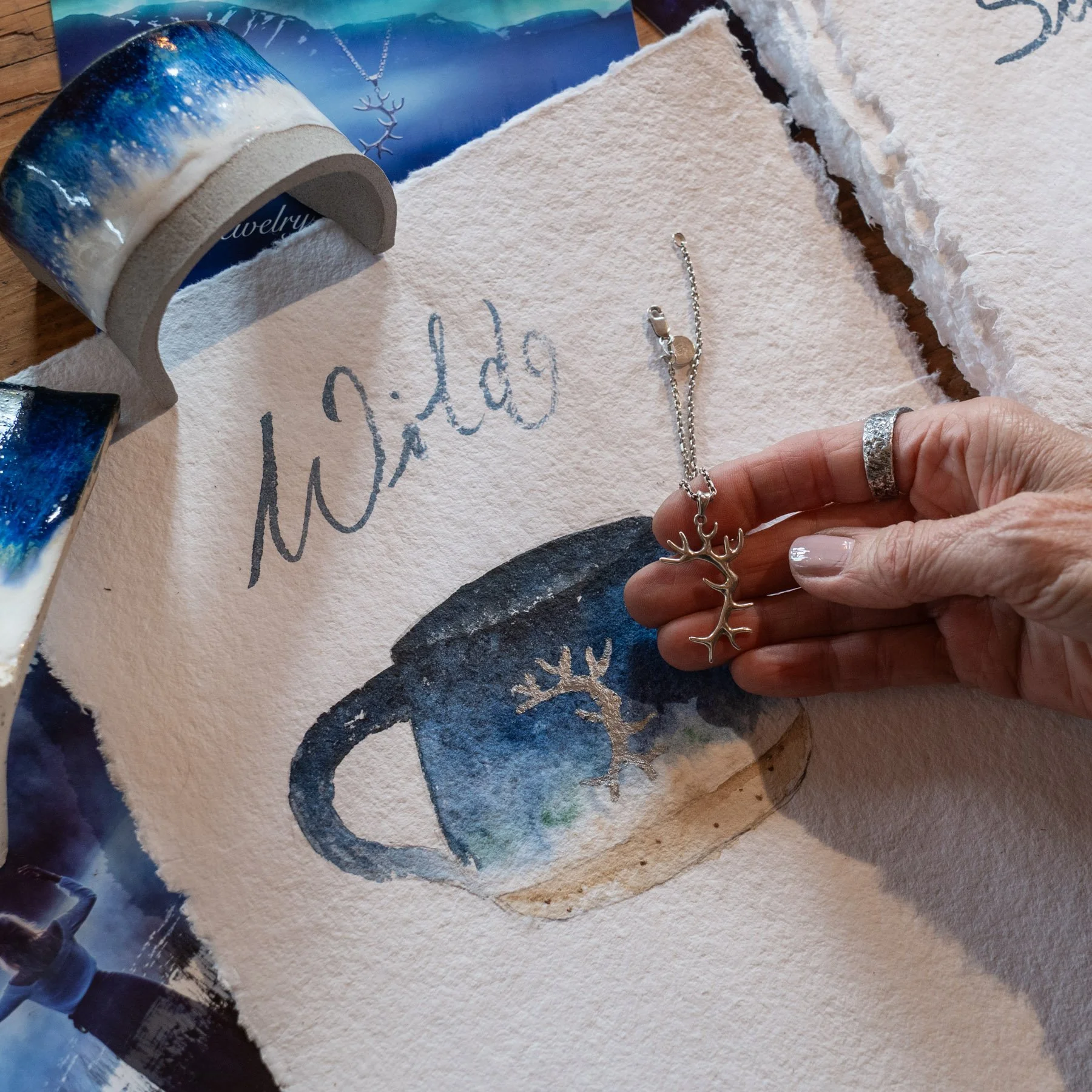 Eine Hand hält eine Halskette mit einem Anhänger in Form eines Rentiers, auf einem Kärtchen mit einem handgemalten Wasserkrug mit einem Rentierkopf-Design. Im Hintergrund sind Wäscheklebebänder mit winterlichem Landschaftsmotiv.