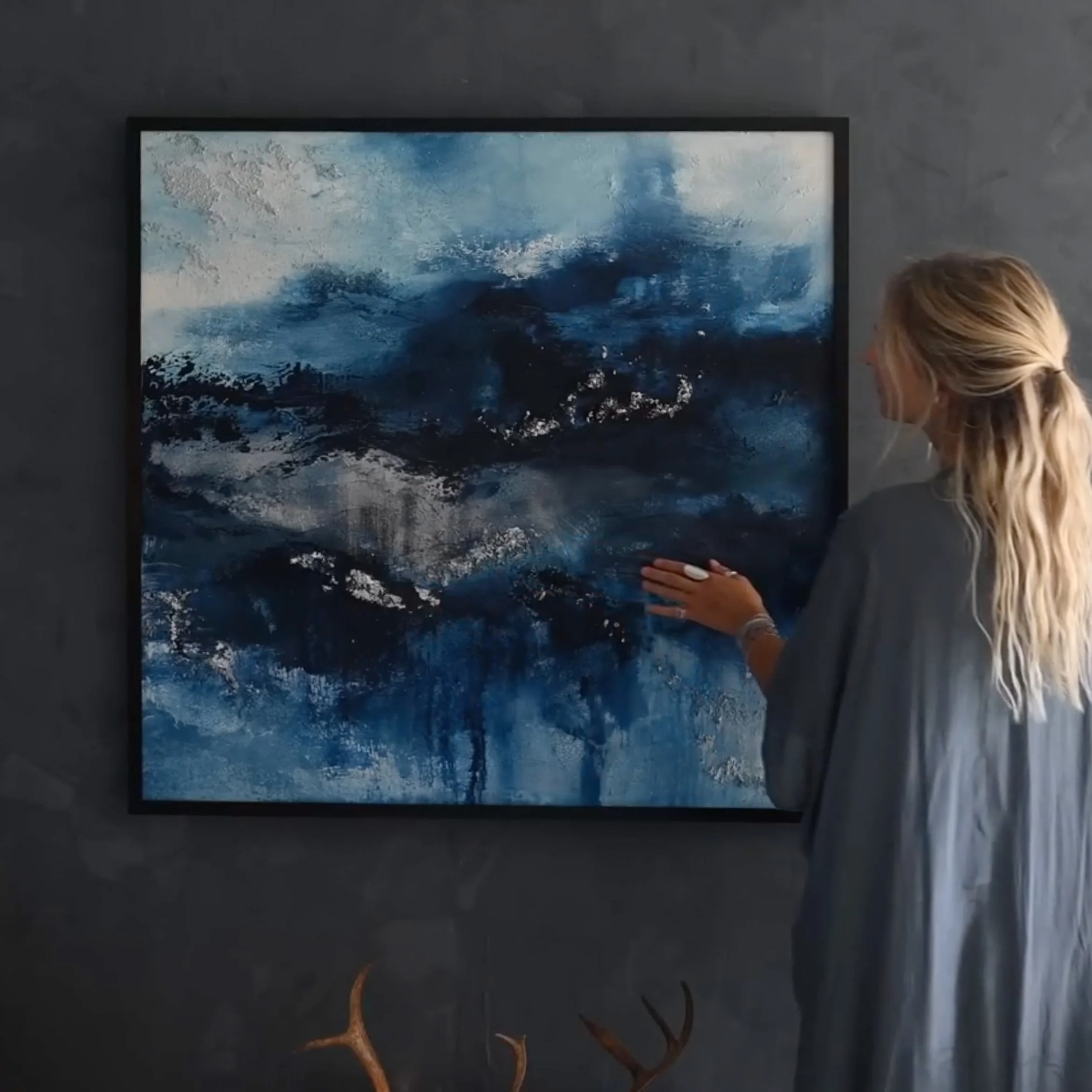 Eine Frau betrachtet ein abstraktes Gemälde mit blauen und weißen Tönen an einer dunklen Wand.