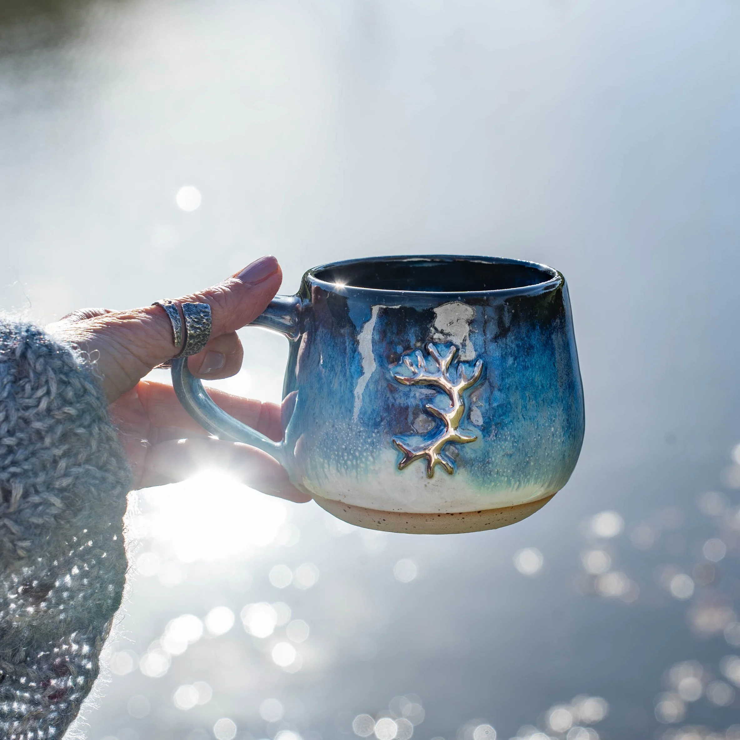 Hand hält einen Keramiktasse mit einem eingefärbten Blauton und einem silbernen Majestät auf der Außenseite, mit Wasser im Hintergrund und Sonnenlichtreflexionen.