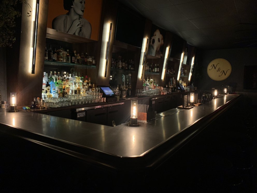 Dimly lit bar interior with a long counter, illuminated liquor shelves, and decorative wall art.