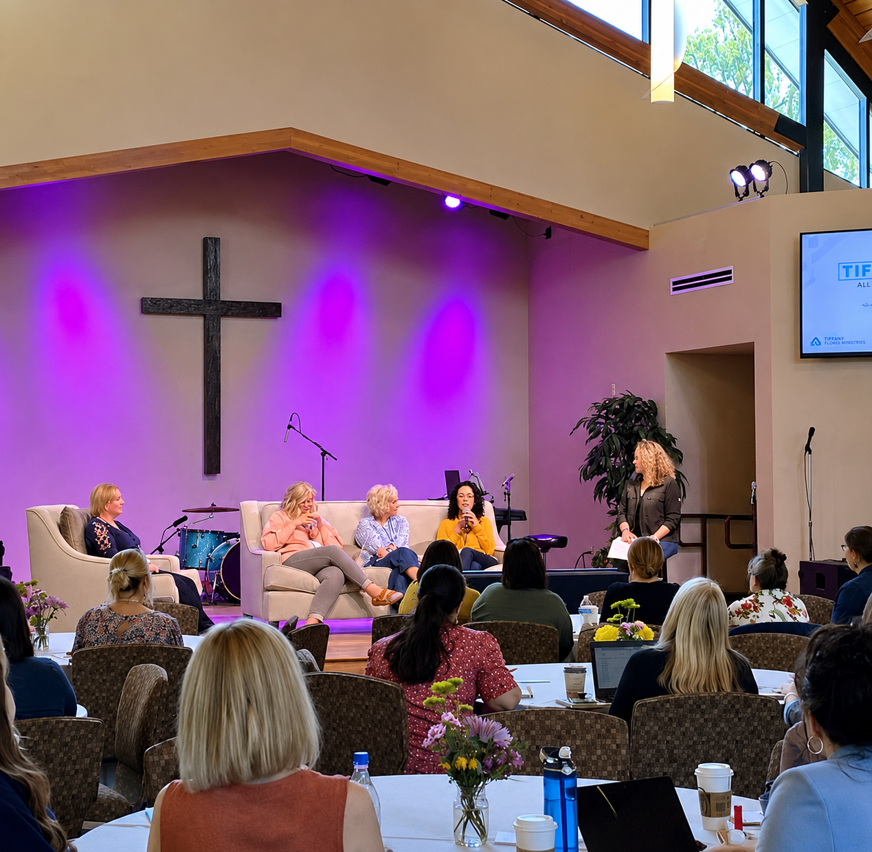 Panel discussion featuring four women speaking on stage at a church event with purple lighting, audience seating, floral table centerpieces, and a large cross displayed behind them.