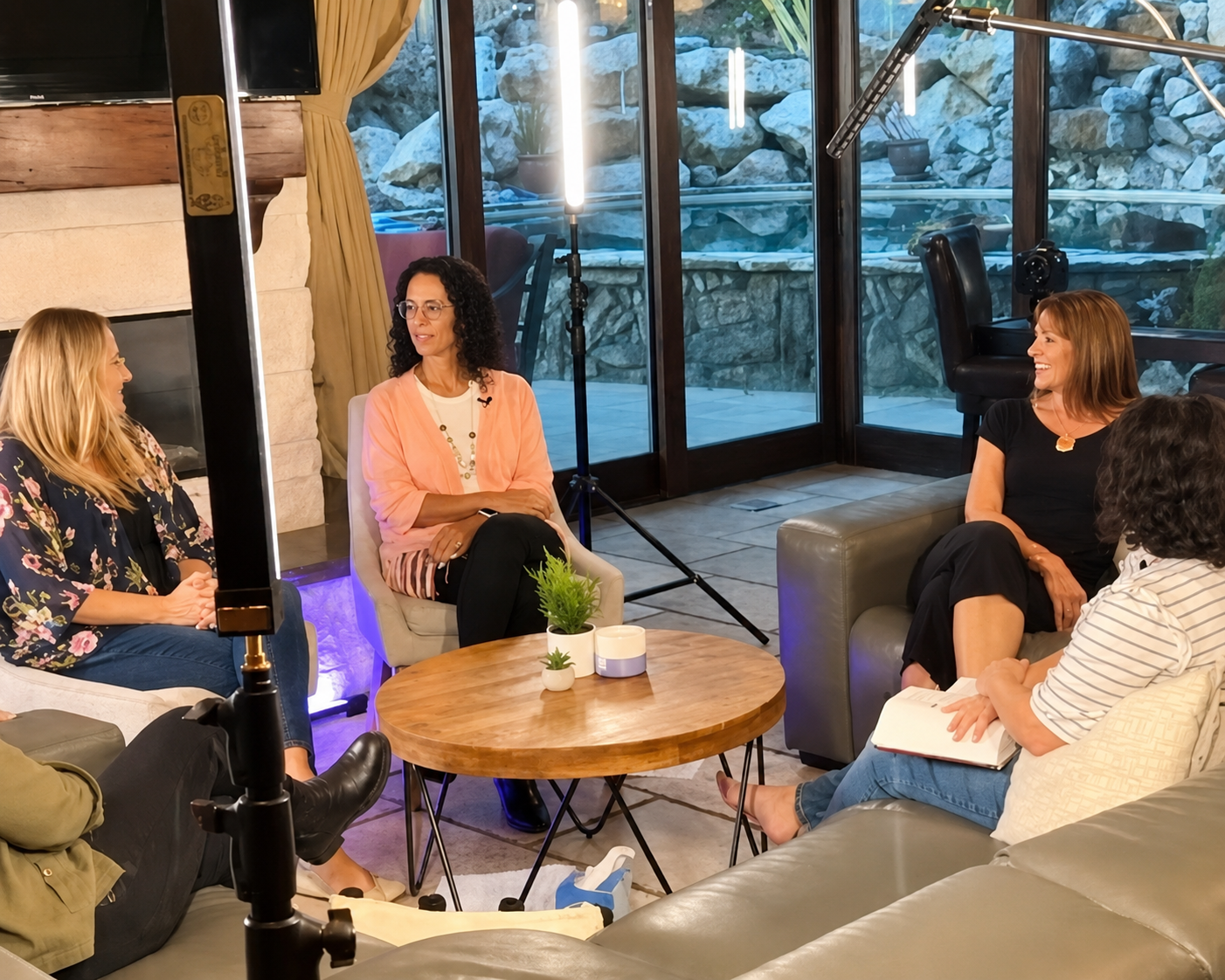 “Women seated in a cozy living-room style discussion circle during a filmed conversation, with studio lighting, microphones, and large windows overlooking an outdoor stone patio.”