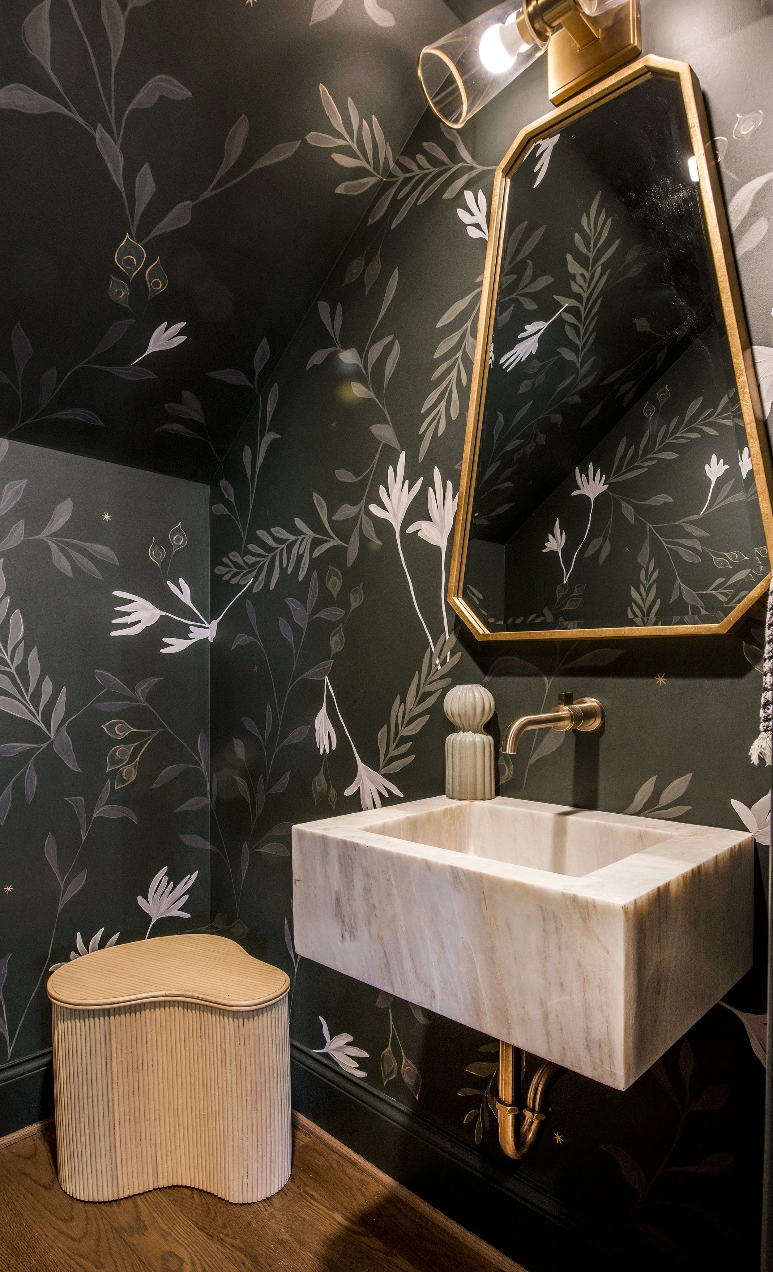 A bathroom with dark floral wallpaper, a marble sink, a gold-framed mirror, a wall-mounted faucet, a cream ceramic vase, and a beige tribute chair.