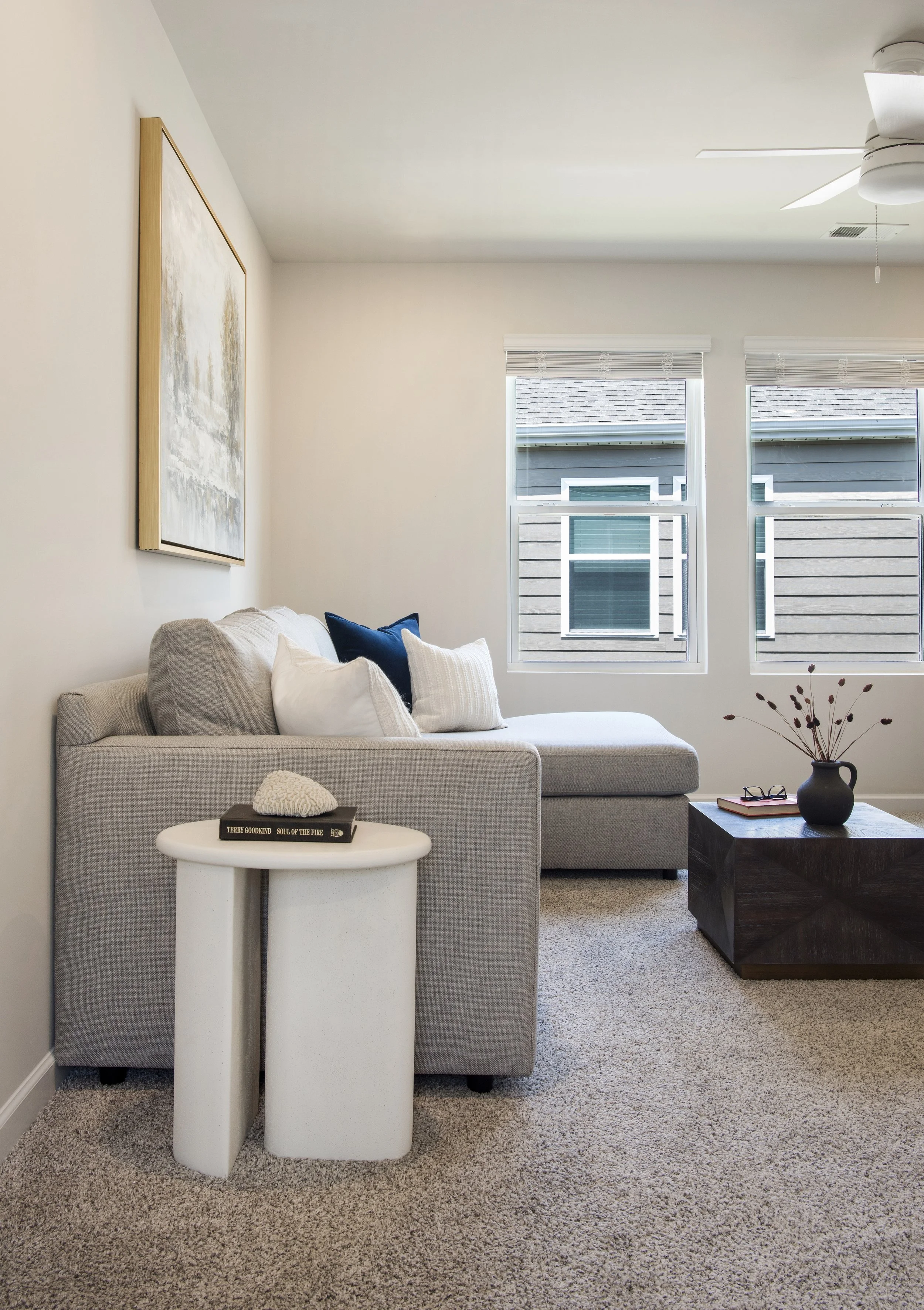 A living room with a light gray sectional sofa with white and dark blue pillows, a small white side table with two books and a decorative object, a dark wooden coffee table with a black vase and dried flowers, two windows with blinds, a ceiling fan, and a piece of abstract wall art.