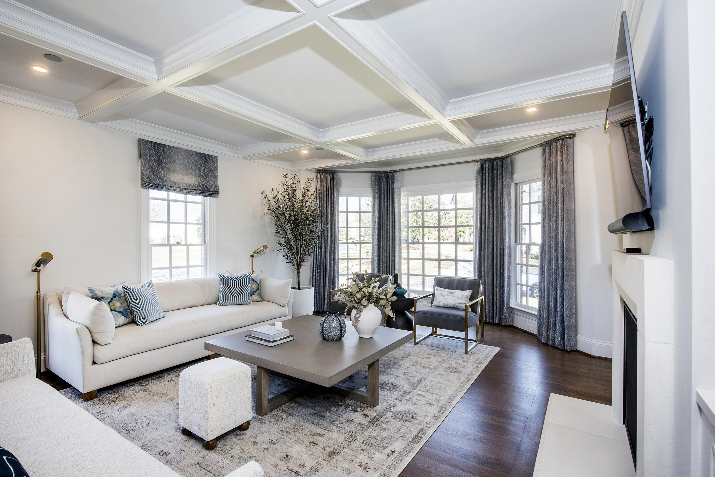 Living room with white walls, large windows with gray and dark curtains, a white sofa with patterned pillows, a rectangular coffee table with decorative items, and a gray armchair. There is a wall-mounted TV near the fireplace, ceiling coffers, hardwood floors, and modern decor.