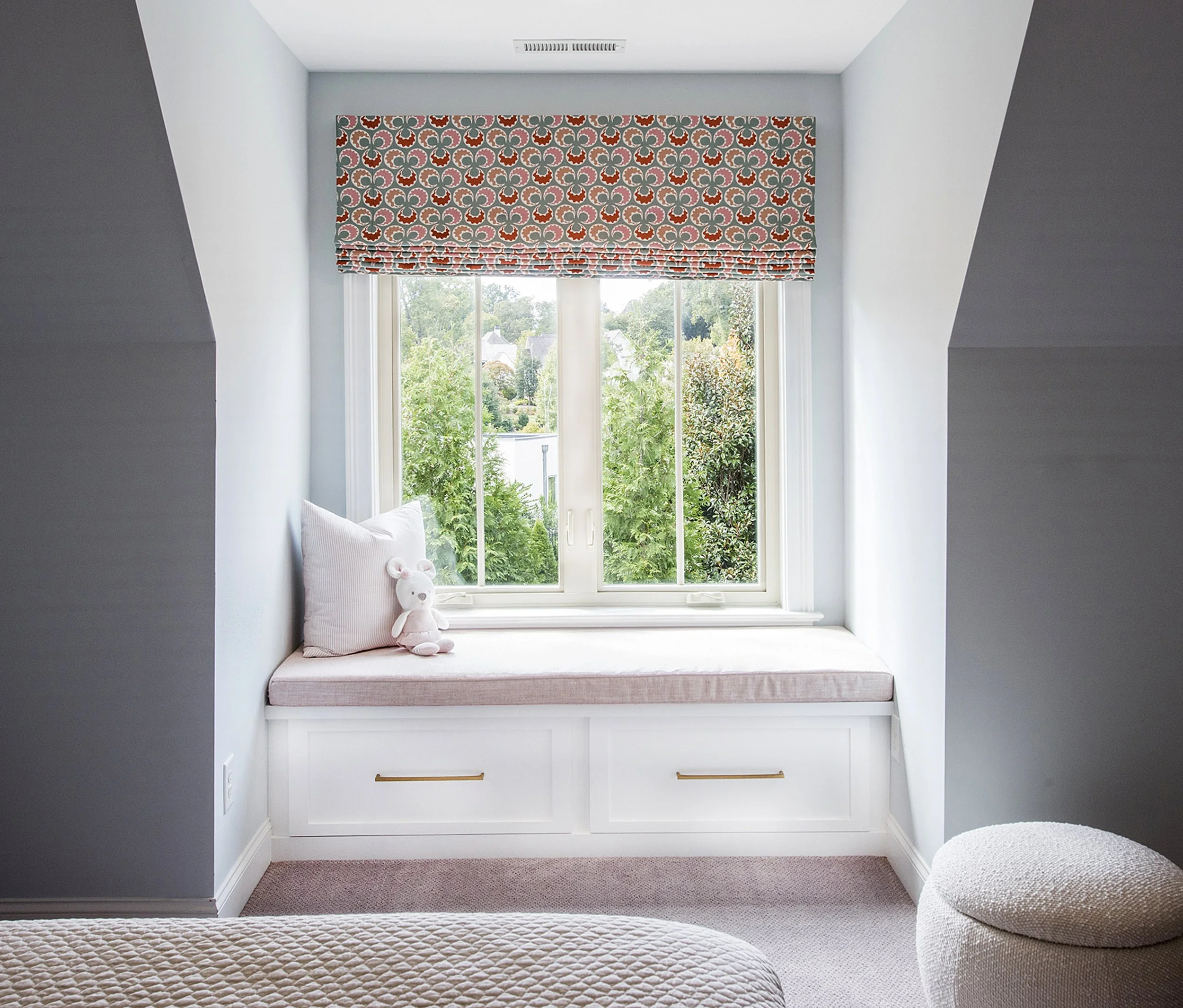 A cozy window nook with a cushioned bench, a pink pillow, a stuffed bear toy, and a window with a colorful patterned valance. Outside view shows greenery and houses.