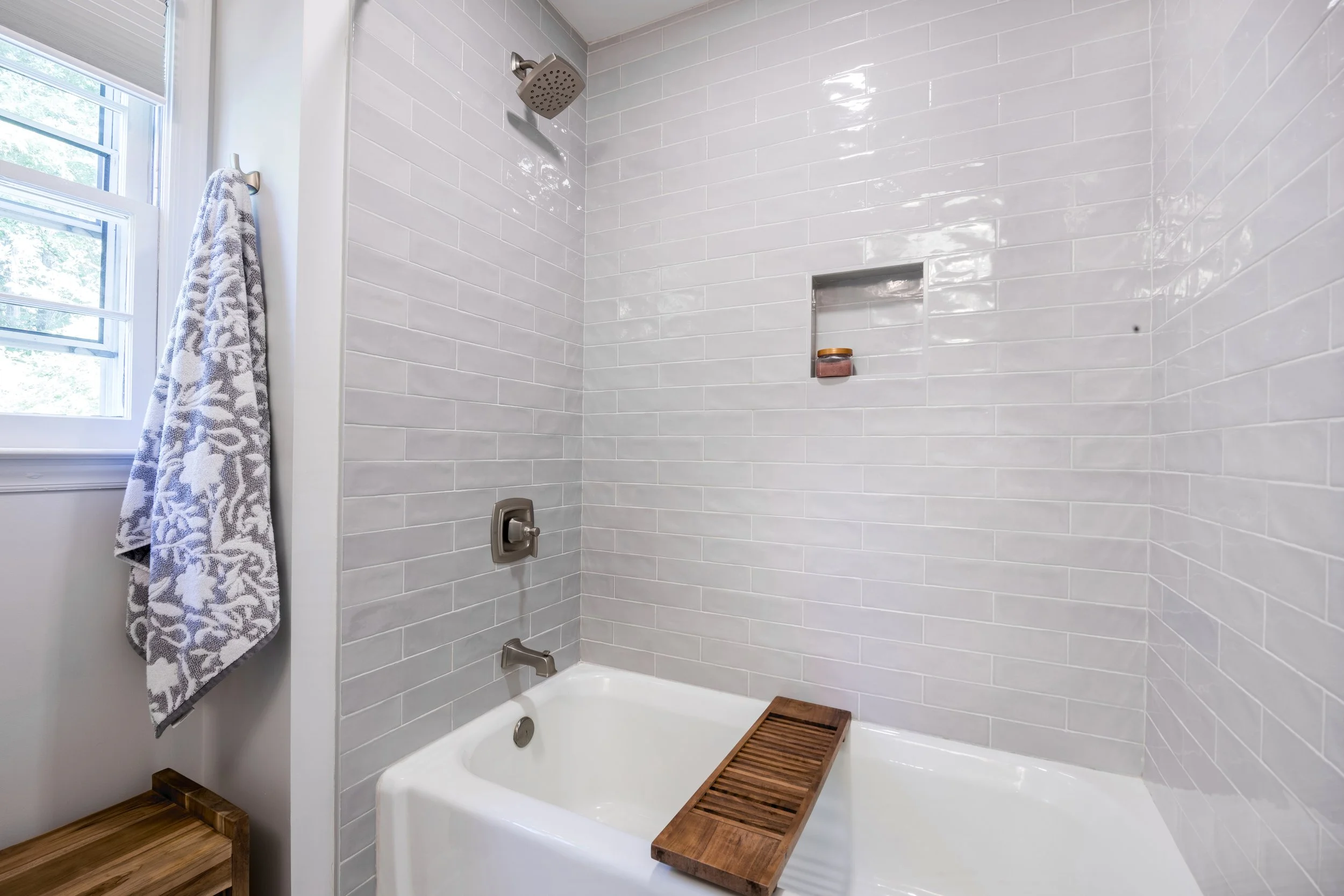 Bathroom shower area with white subway tile walls, a ceiling shower head, a built-in recessed shelf with a soap jar, a wall-mounted faucet, and a wooden bath tray, near a window with a patterned towel hanging on a hook.