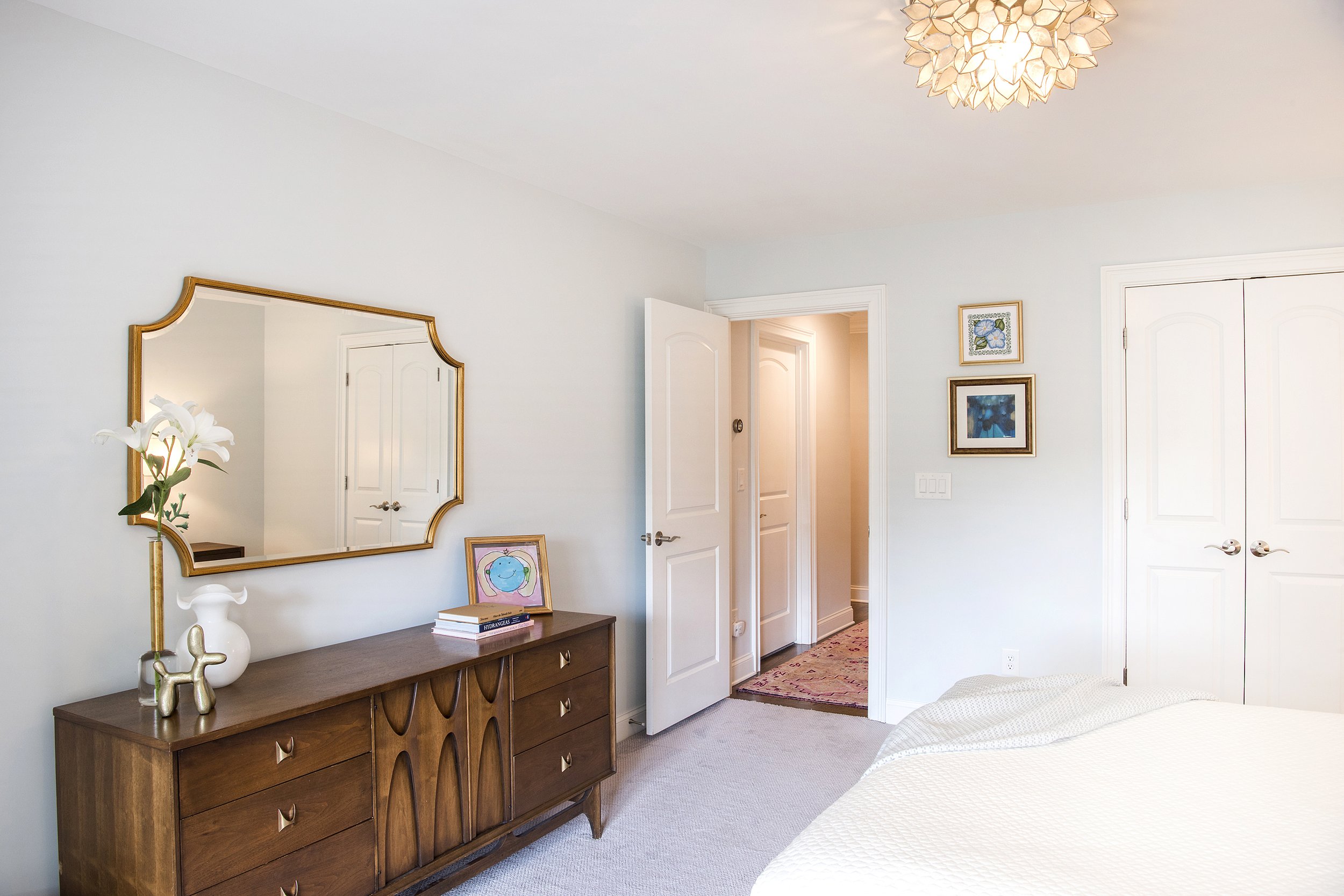 A bedroom with a wooden dresser, a large mirror, a vase with a white lily, framed artworks, a bed covered with a white blanket, and a closed closet door. The room has light-colored walls and a ceiling light fixture.